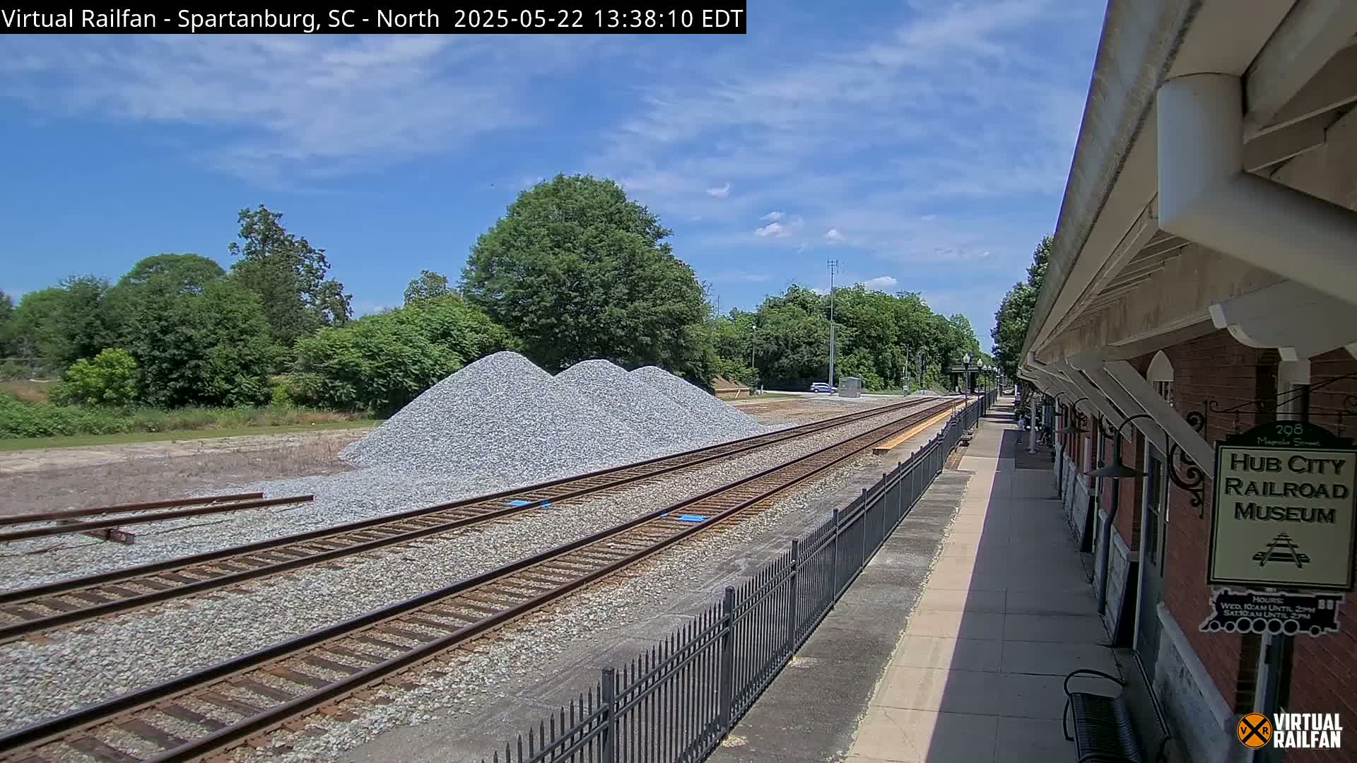 Train tracks, bordered by a black metal fence, extend from a brick railroad museum toward piles of gravel under a partly cloudy sky.