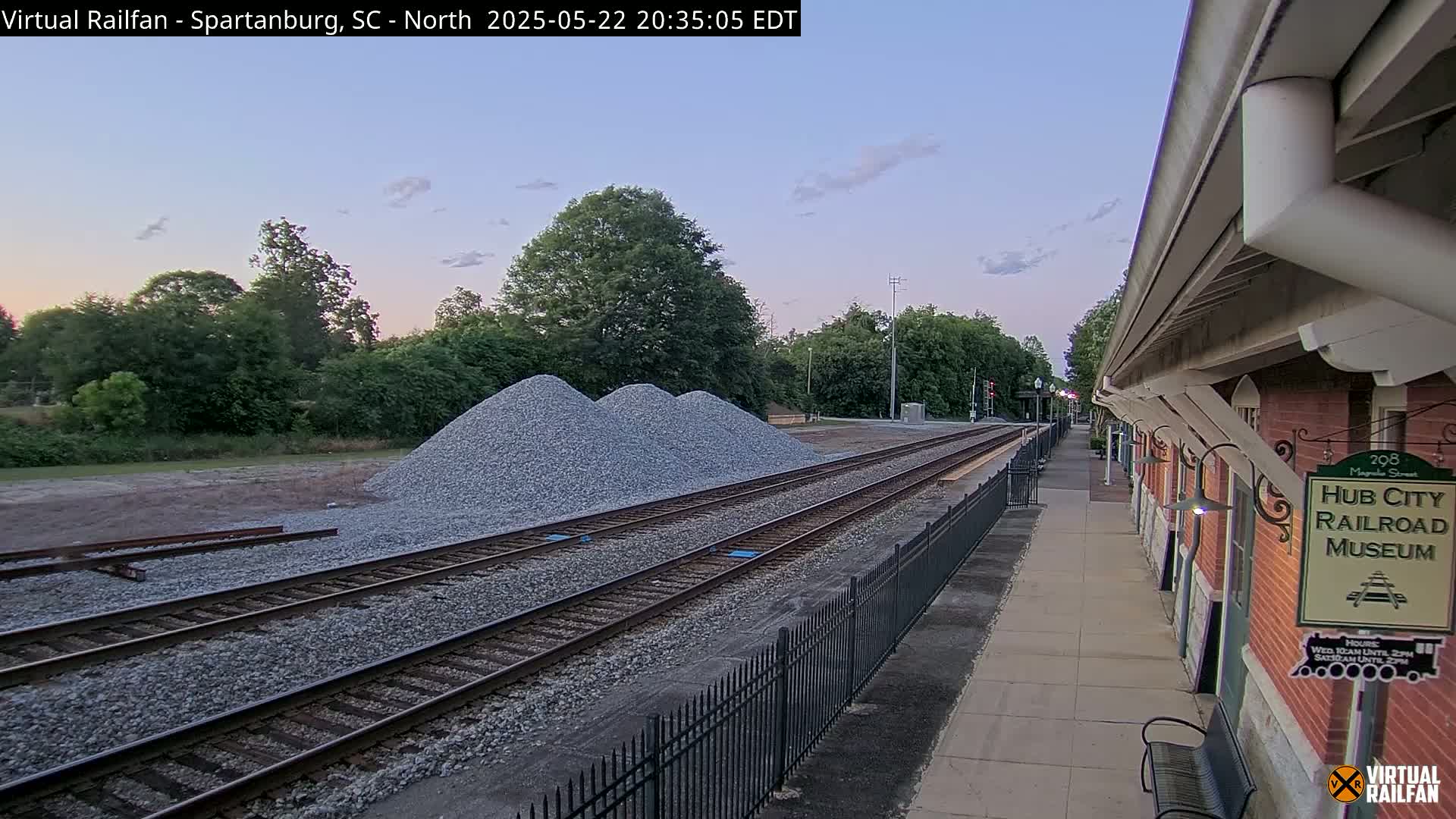 A railroad track with piles of gravel next to it runs alongside a brick railroad museum under a twilight sky.
