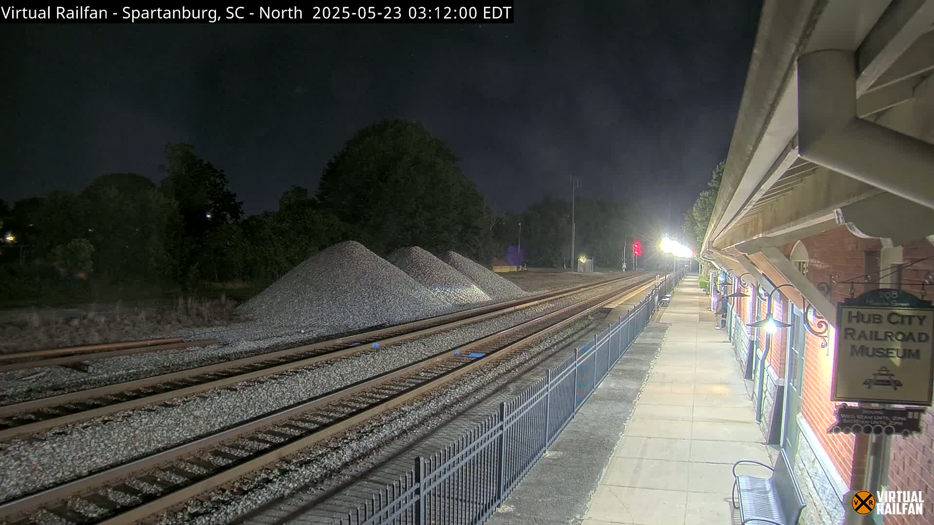At night, railroad tracks extend toward piles of gravel next to a brick train station platform.
