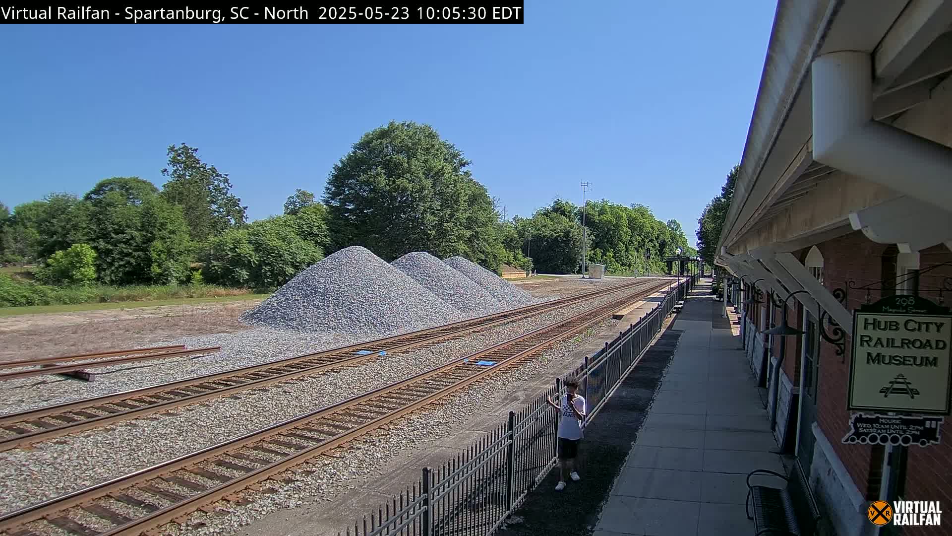 Under a clear blue sky, railroad tracks extend past piles of gravel toward a brick building and lush green trees.