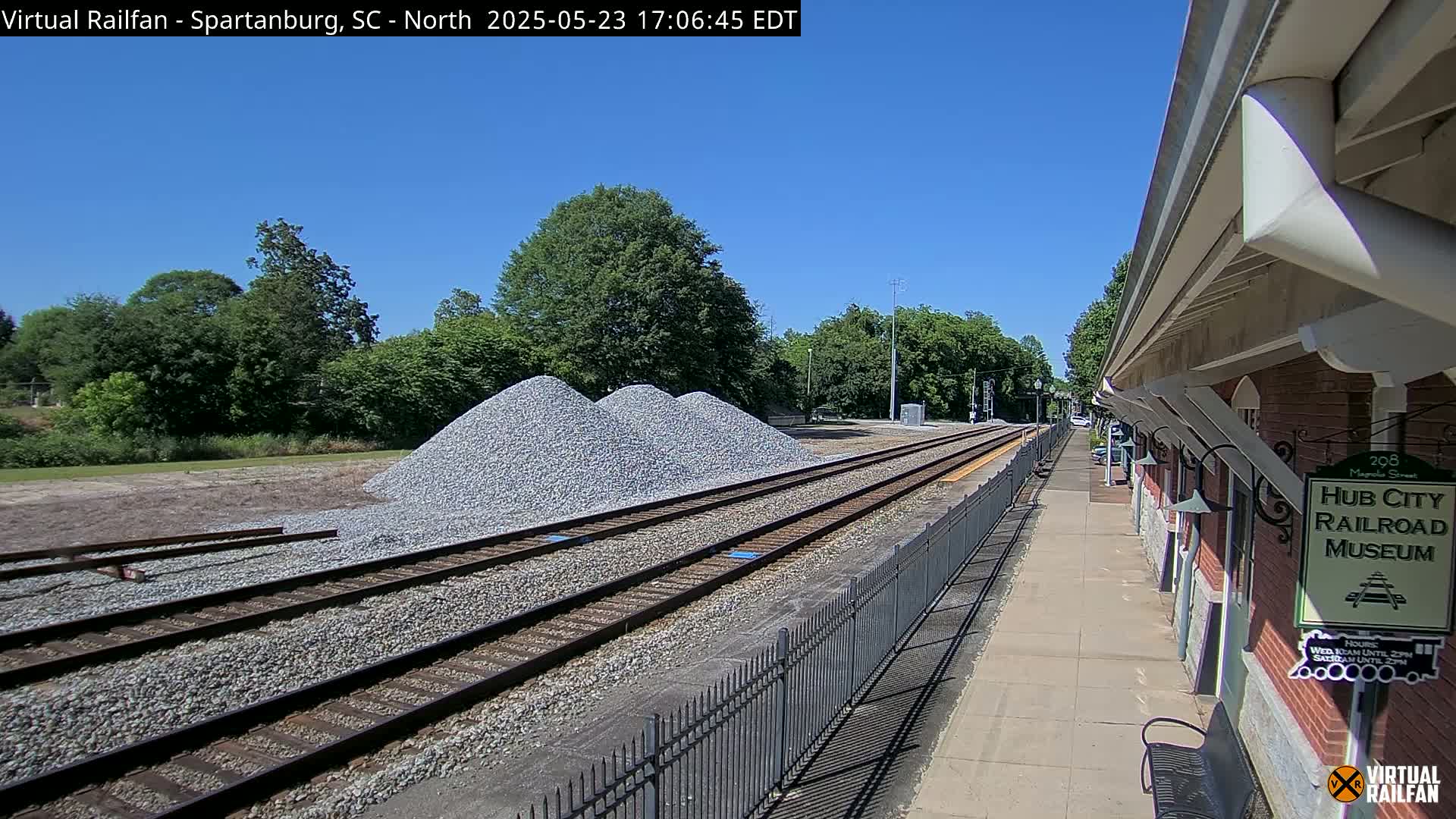 Under a clear blue sky, railroad tracks extend from a train depot past piles of gravel toward a line of trees.