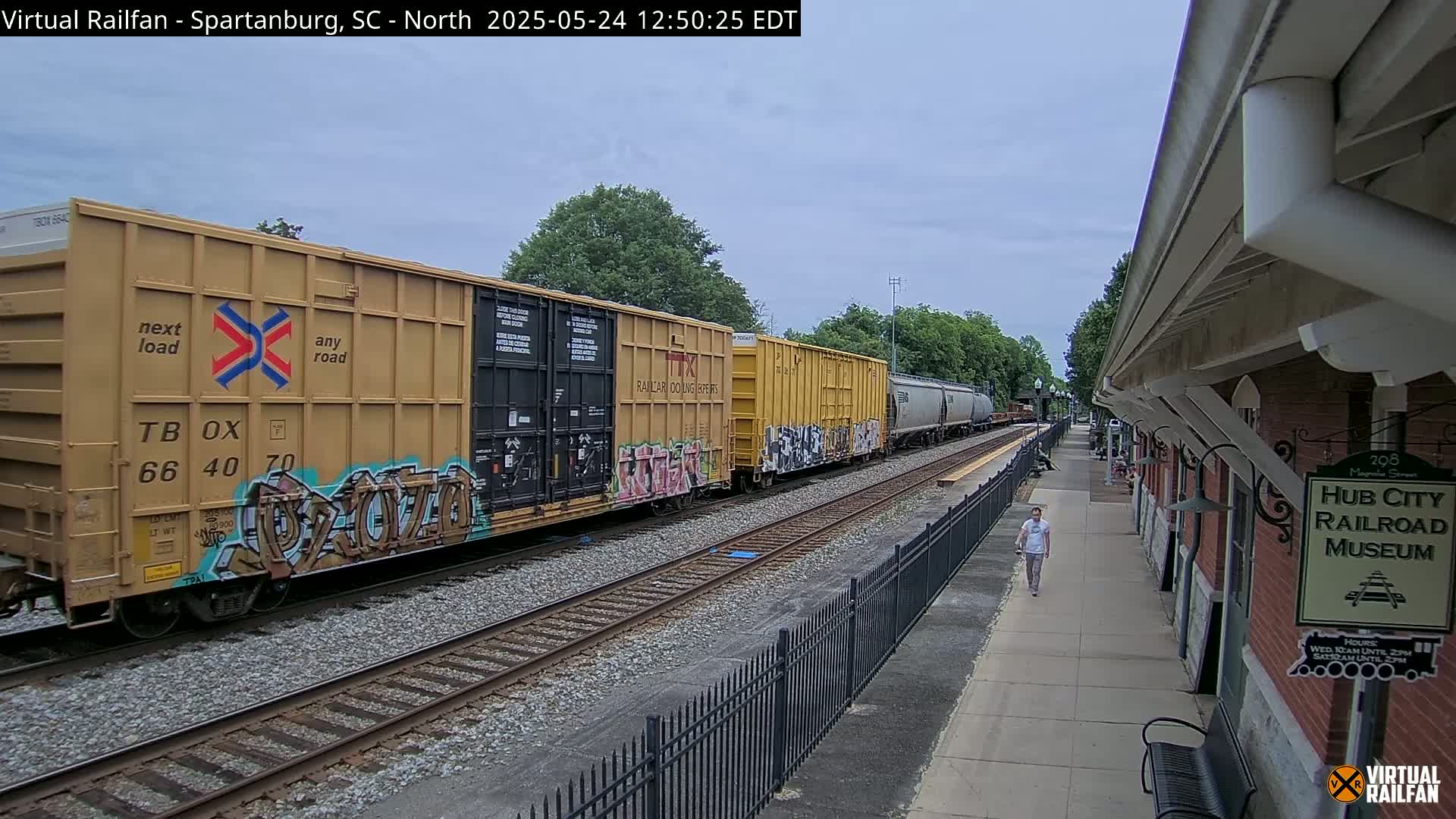 A freight train with graffiti-covered cars passes a brick railroad museum building on a partly cloudy day.