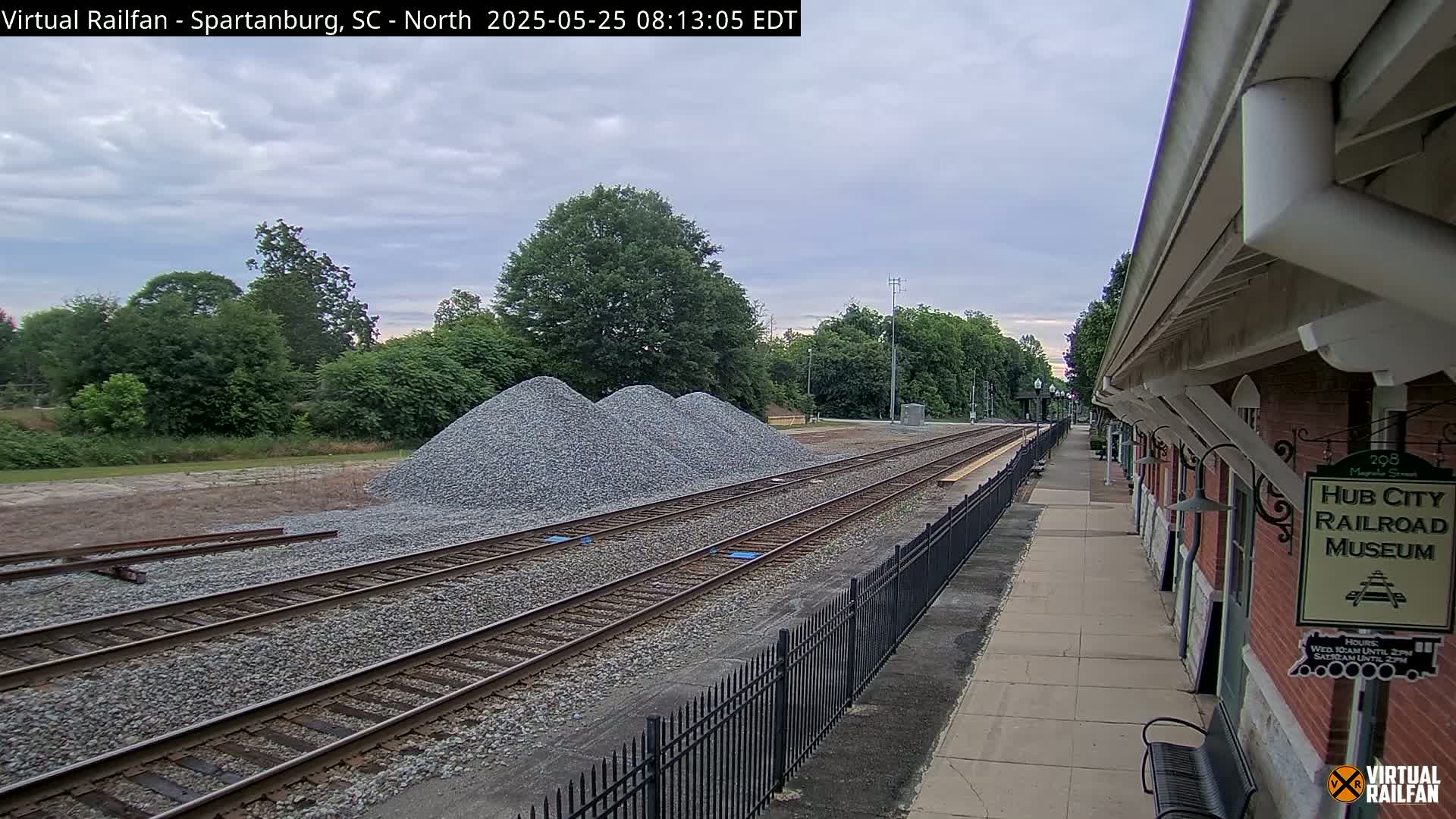 Several piles of gravel sit beside railroad tracks next to a brick railroad museum under an overcast sky.