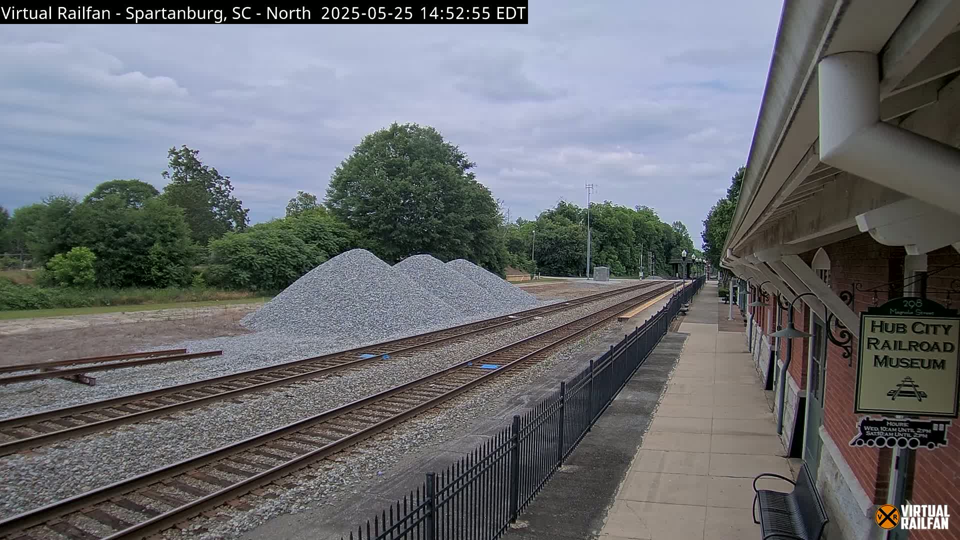 Three piles of gravel sit beside a train track next to a brick building, under a partly cloudy sky.