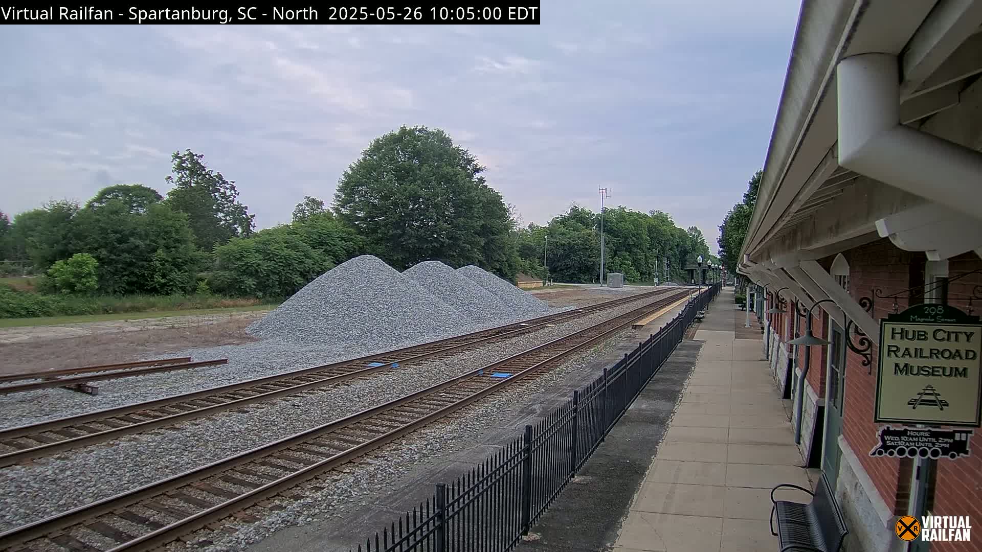 Under an overcast sky, railroad tracks extend from a brick building alongside a paved walkway, with piles of gravel situated nearby.