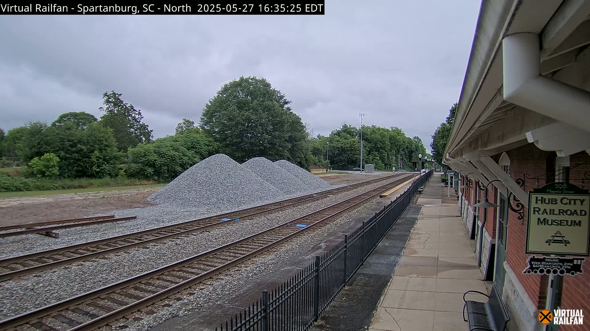 Under an overcast sky, railroad tracks extend from a train platform past several piles of gravel toward a line of trees.
