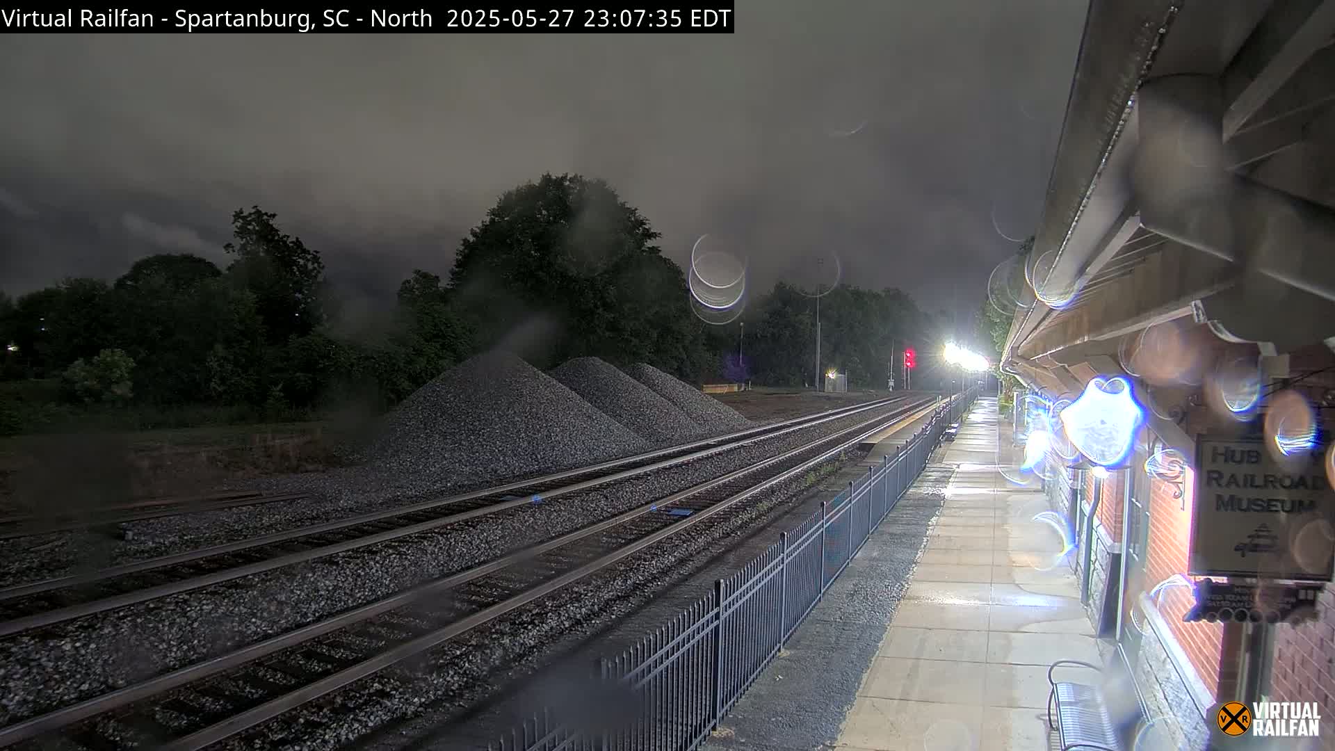 A nighttime view of rain-slicked train tracks, bordered by a fence and a brick building, with piles of gravel nearby under a dark, overcast sky.
