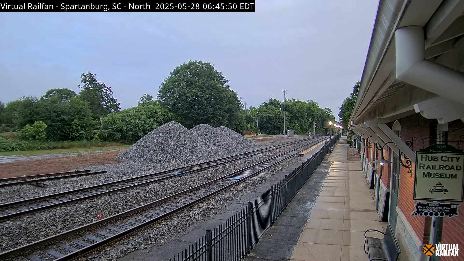 Several piles of gravel are situated beside railroad tracks next to a brick railroad museum building under an overcast sky.
