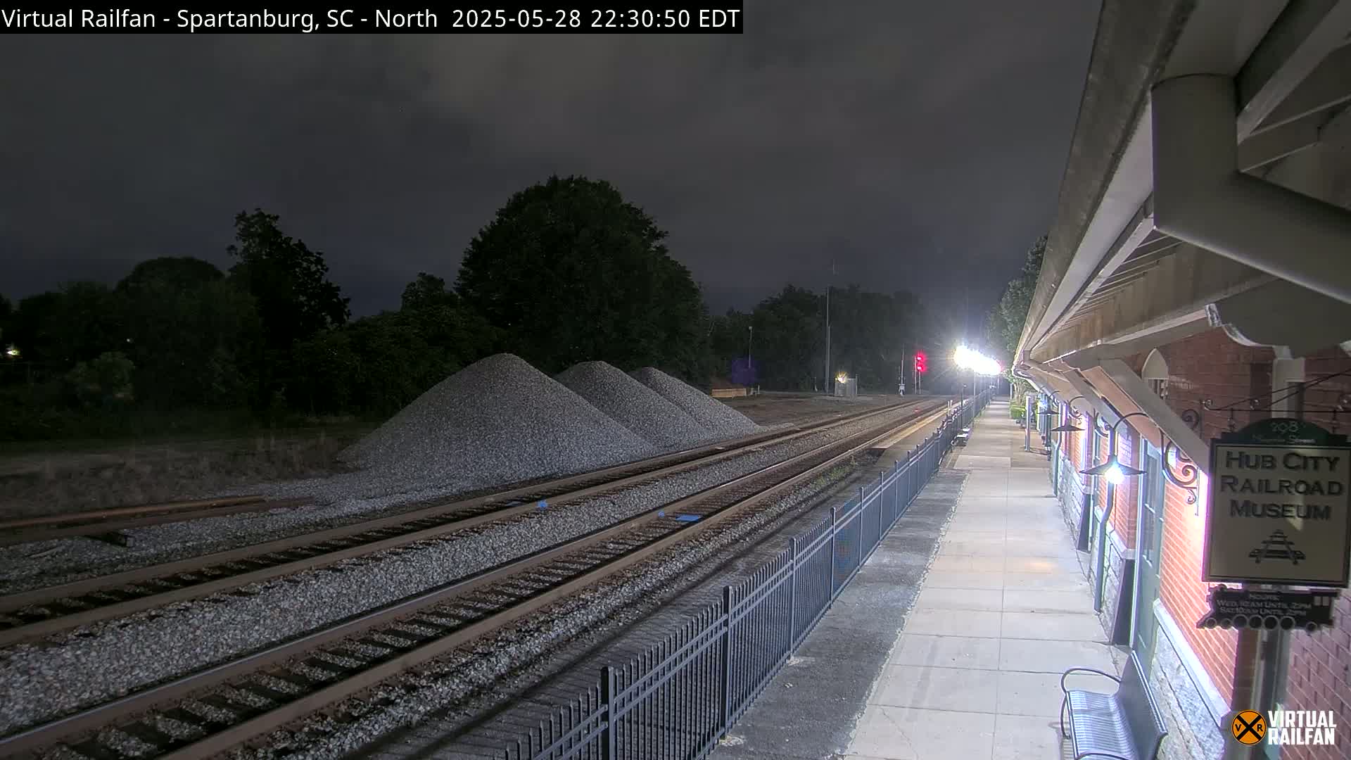 Under a dark, nighttime sky, railroad tracks extend from a train platform next to a brick building toward several piles of gravel.