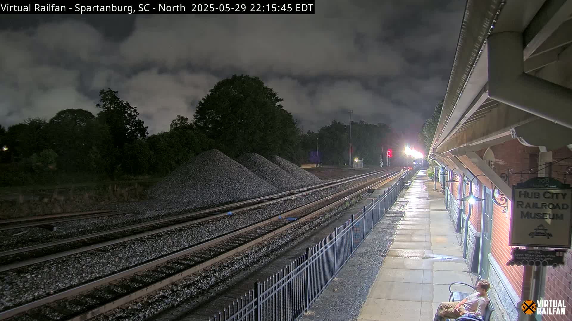 At night under a cloudy sky, railroad tracks extend from a train platform next to a brick building towards piles of gravel.
