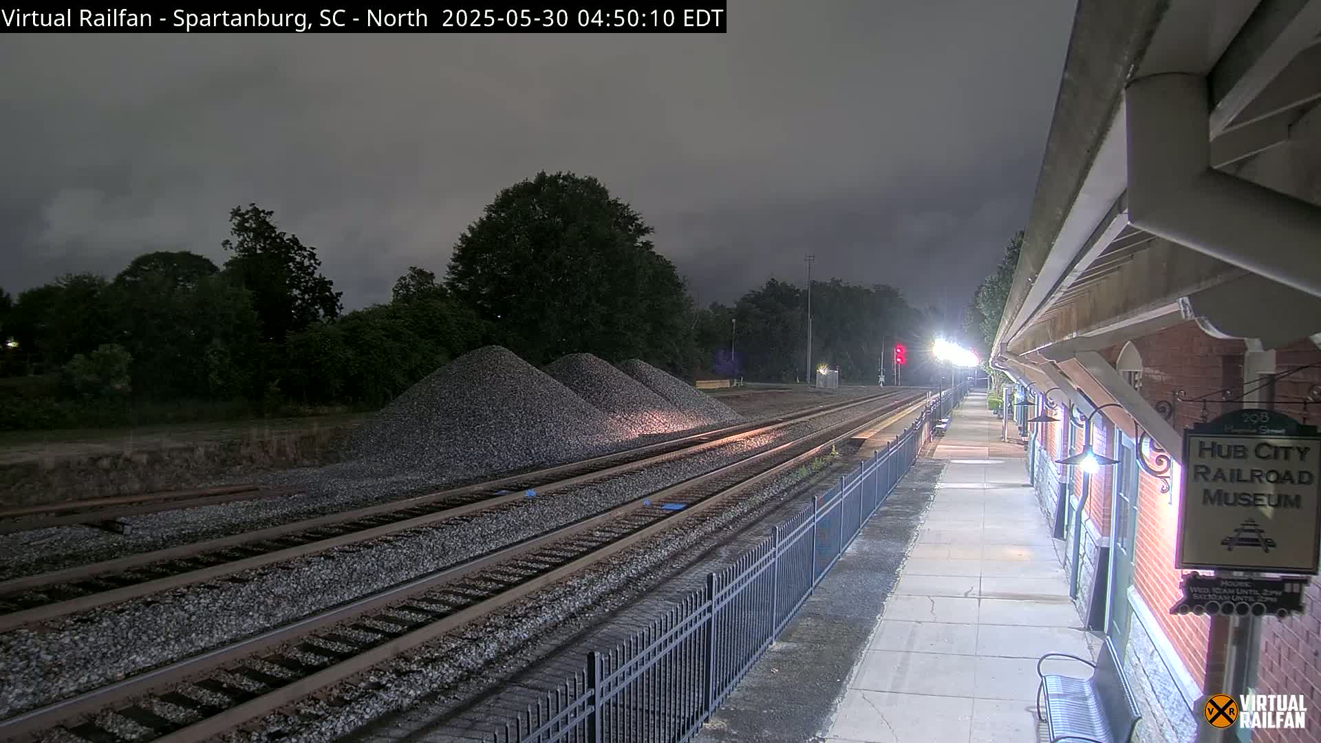 Under a dark, overcast night sky, a railway station platform extends alongside train tracks, with piles of gravel nearby.