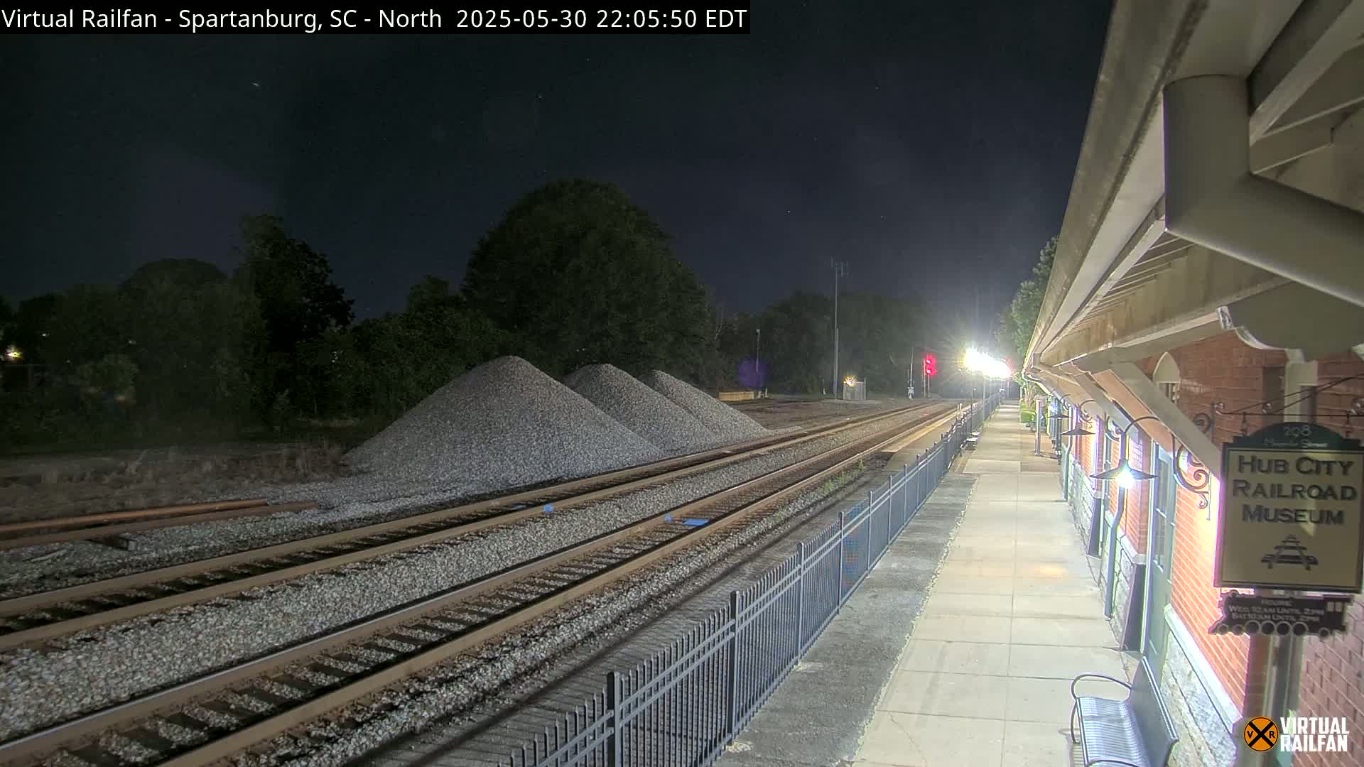 At night, a train station platform next to multiple train tracks is illuminated, with piles of gravel nearby.