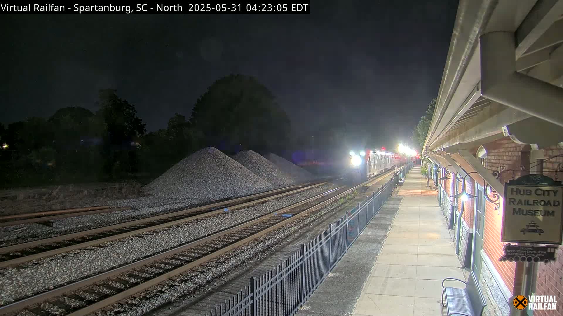 At night, a train approaches a station next to a brick building and piles of gravel.