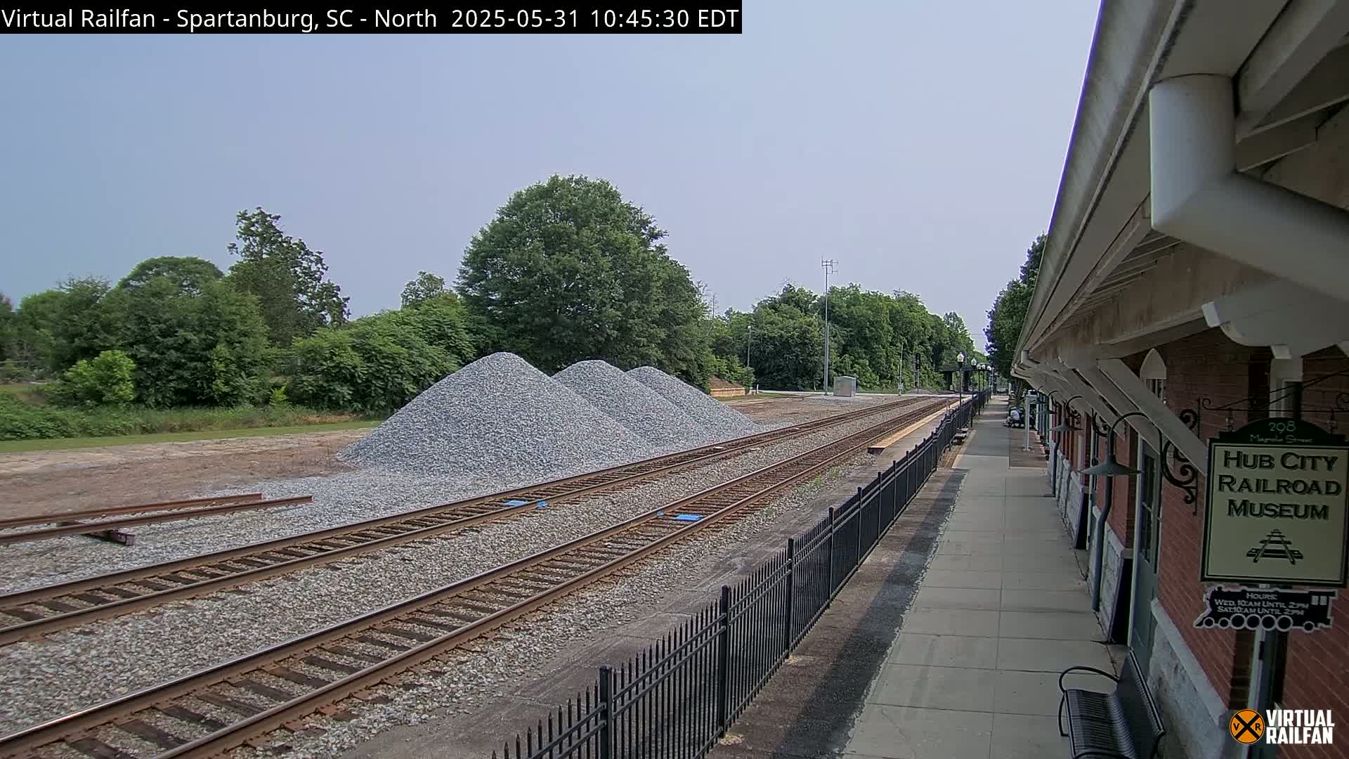 Three piles of gravel sit beside a railroad track next to a brick building, under a partly cloudy sky.