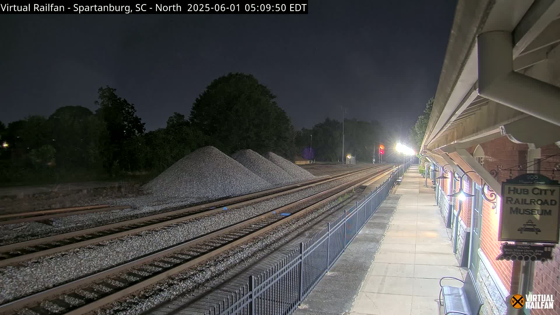 At night, train tracks with several piles of gravel next to them run alongside a lit platform with a brick building.