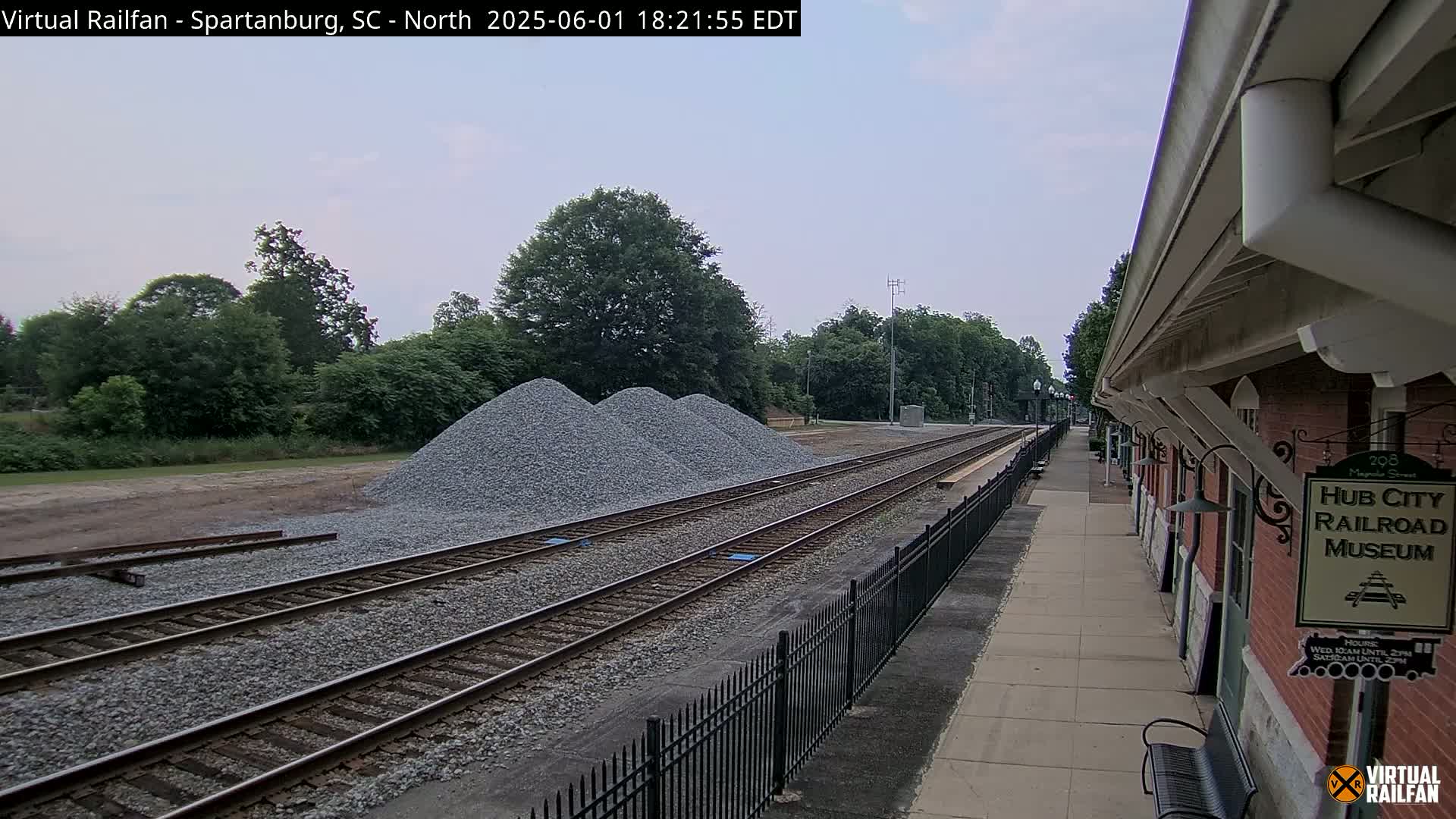 Under a cloudy sky, railroad tracks extend from a train platform beside a brick building toward piles of gravel and a line of trees.