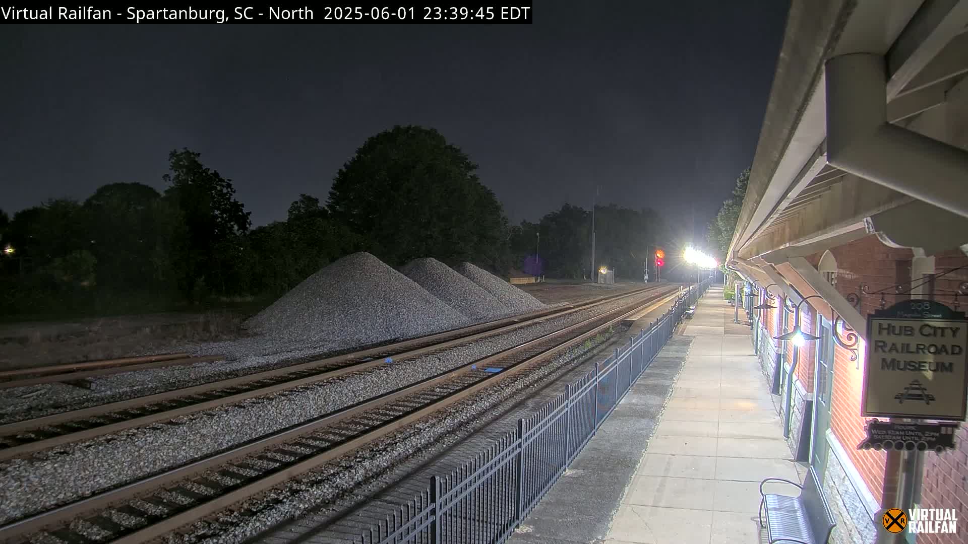 At night, railroad tracks extend from a platform beside a brick building towards piles of gravel under a dark sky.