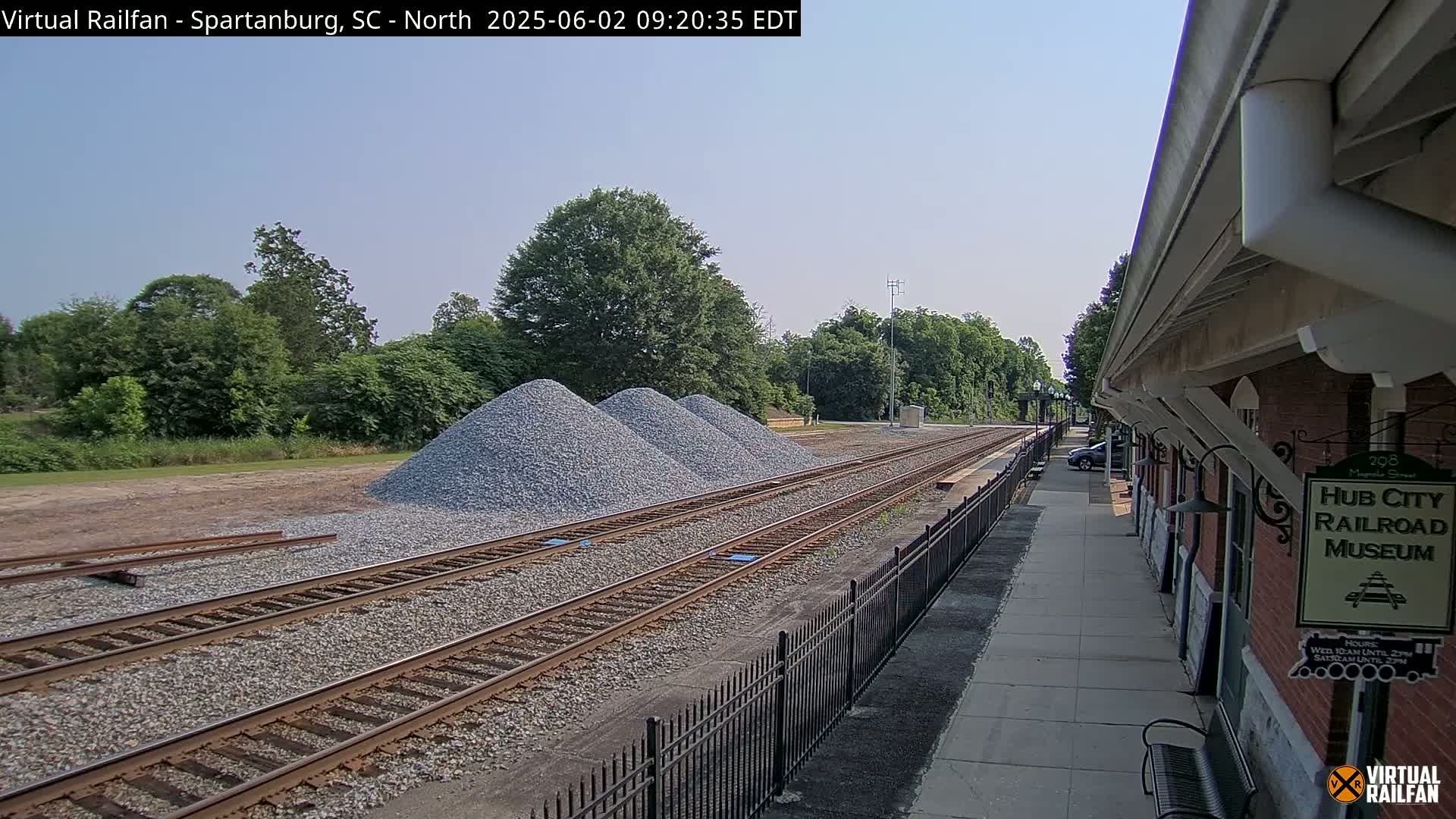 Under a clear sky, several piles of gravel sit beside railroad tracks next to a train platform.