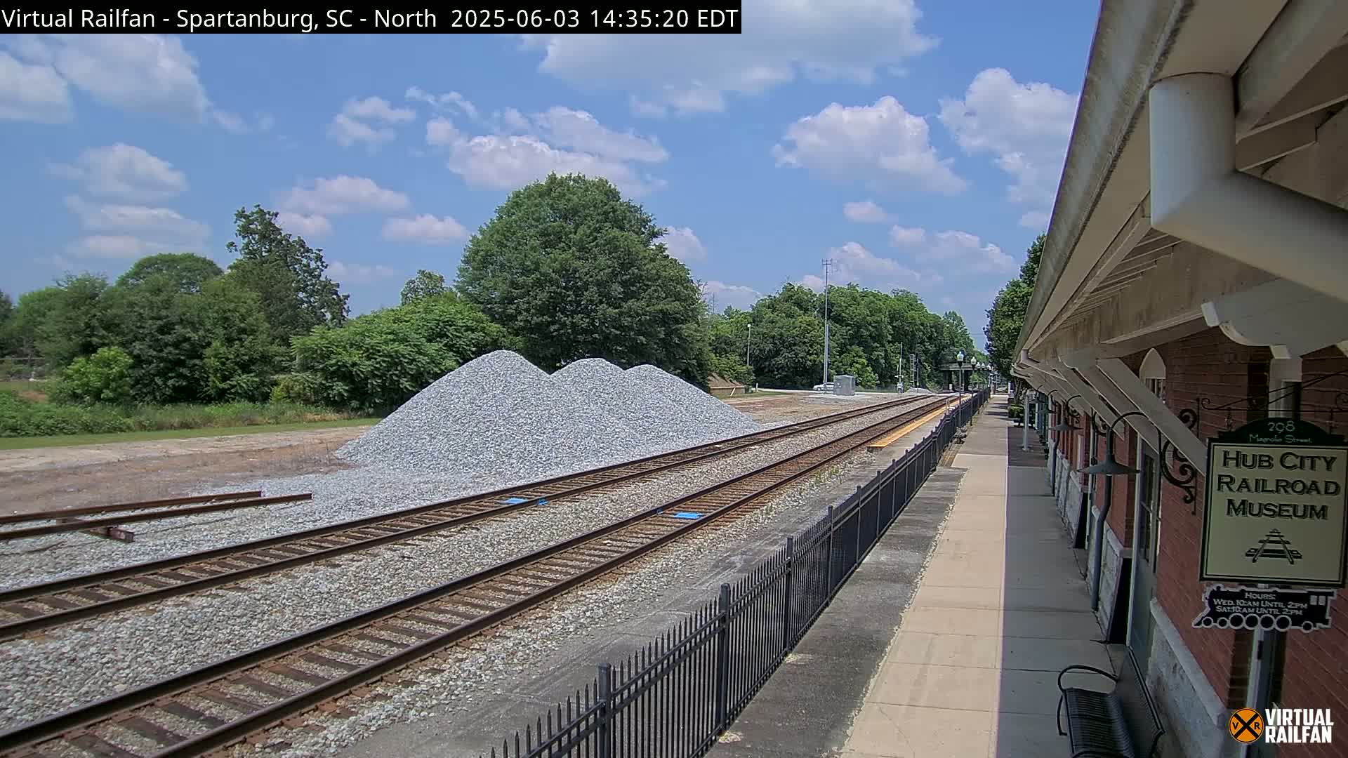 Under a partly cloudy sky, railroad tracks extend from a train platform beside piles of gravel toward a line of trees.