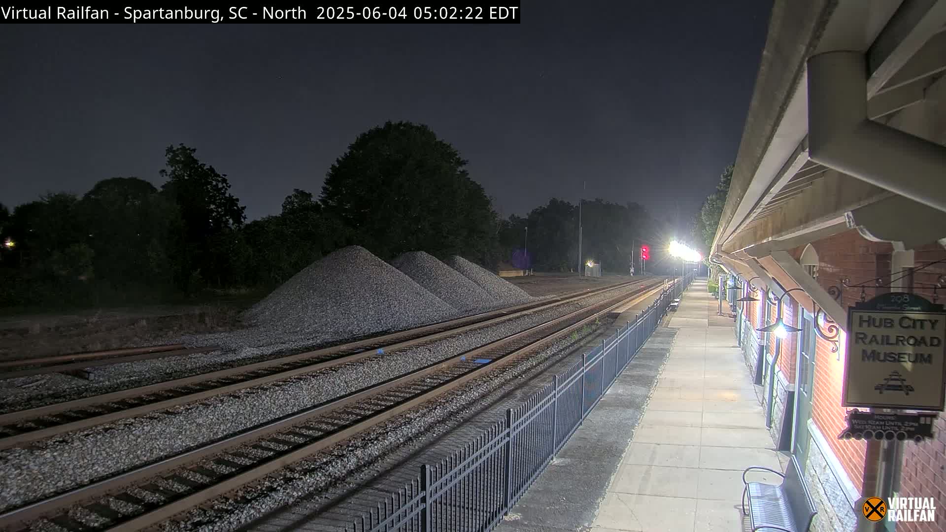 At night, a train station platform is shown with railroad tracks extending into the distance, piles of gravel nearby, and trees in the background under a dark sky.