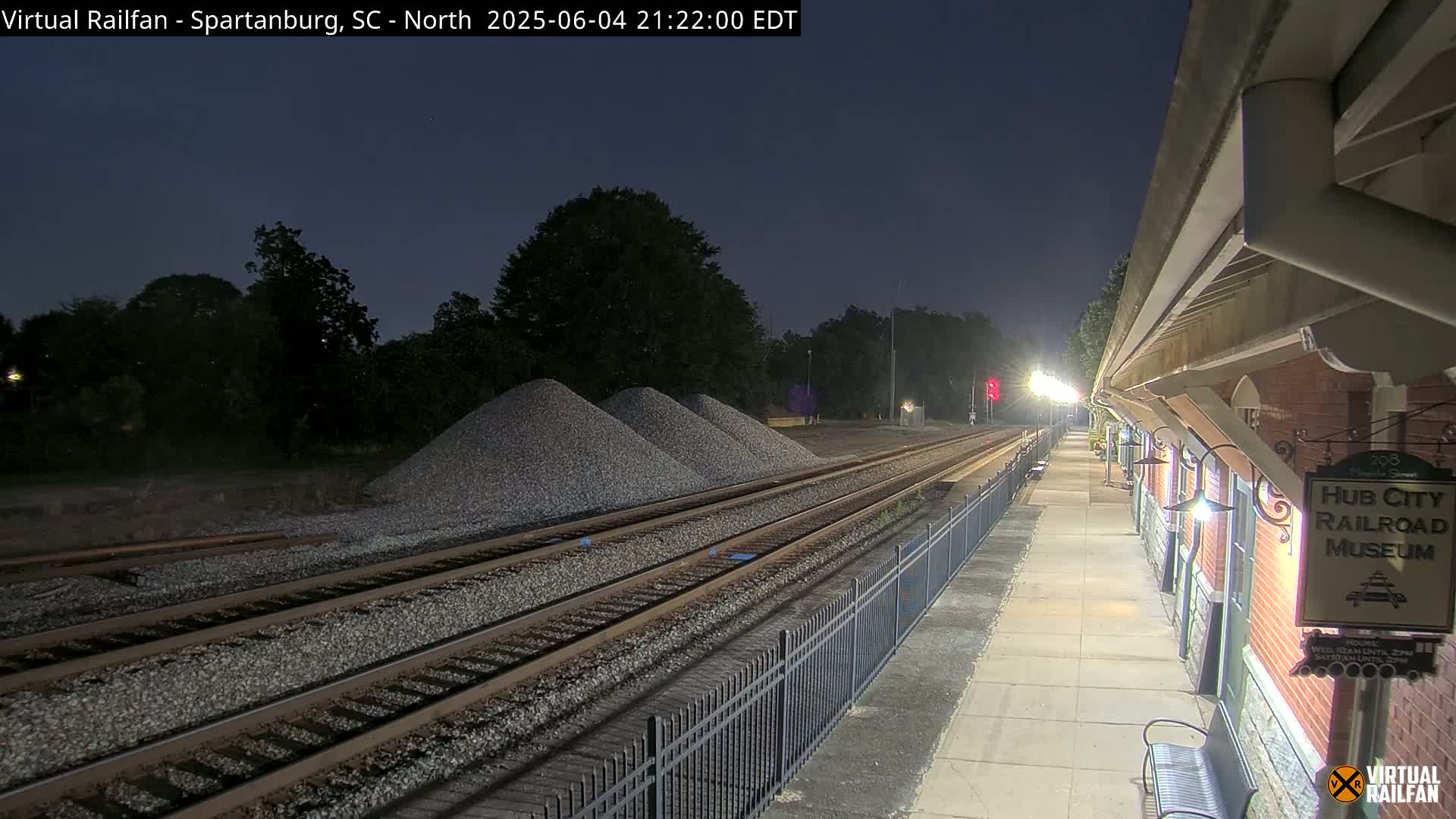 Under a dark night sky, railroad tracks extend from a nighttime train platform toward piles of gravel beside a brick building.