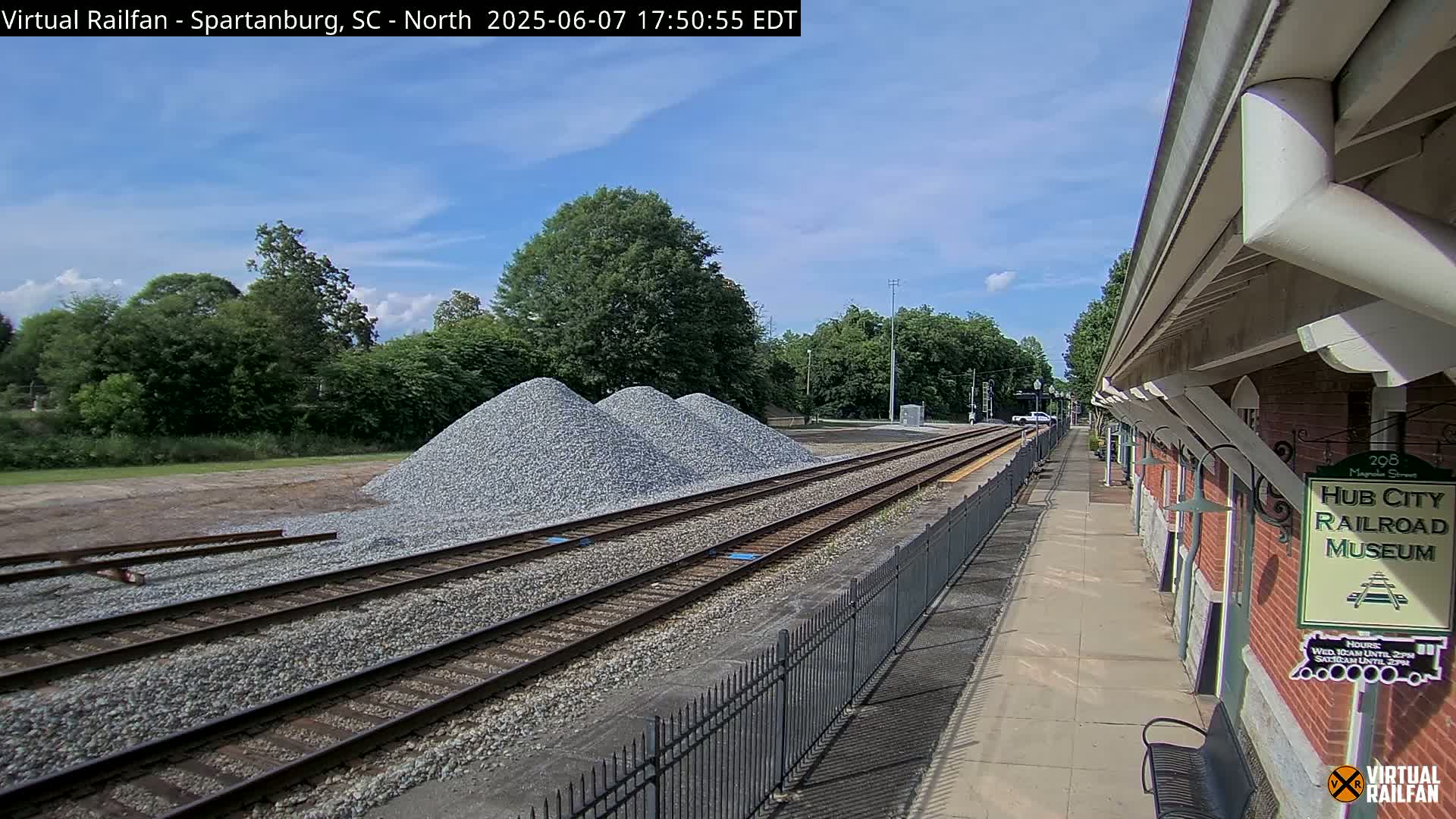 A sunny day shows railroad tracks with piles of gravel beside them, adjacent to a railroad museum platform.