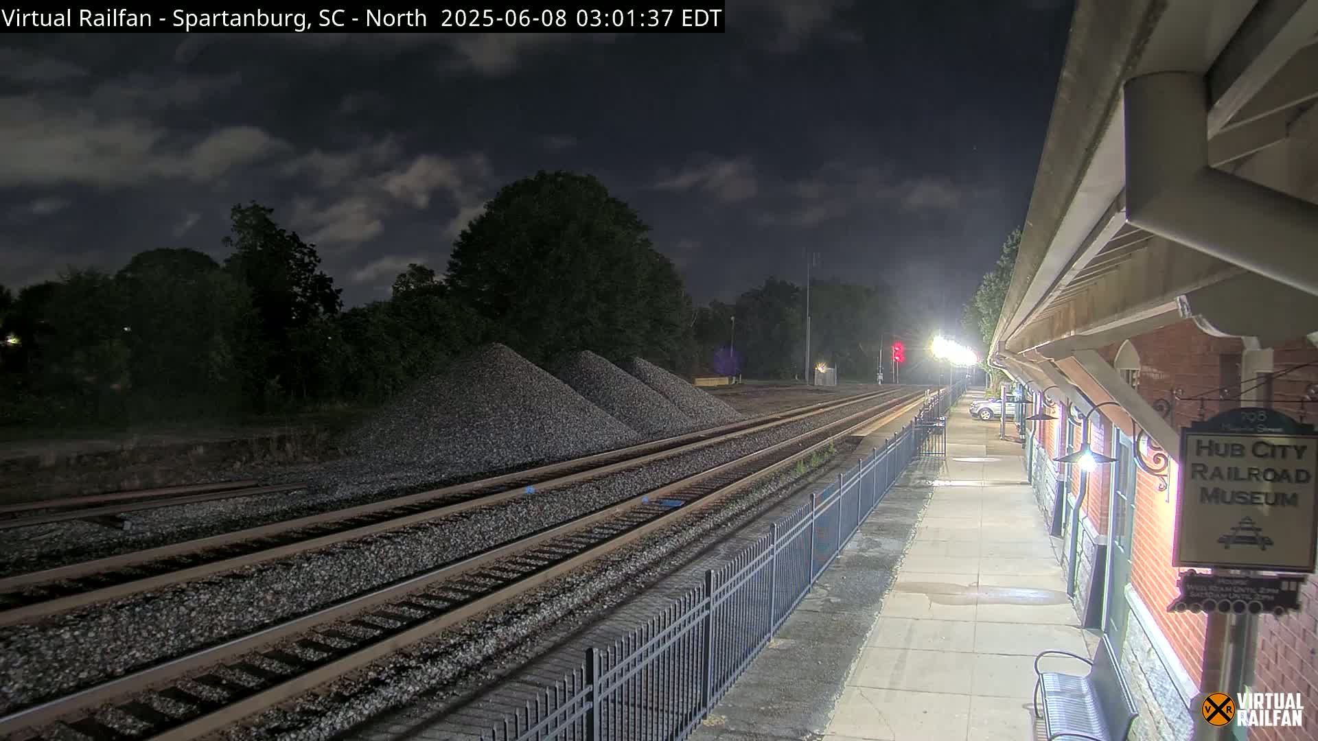 Under a partly cloudy night sky, train tracks extend into the distance past piles of gravel, next to a railroad museum platform with a bench.