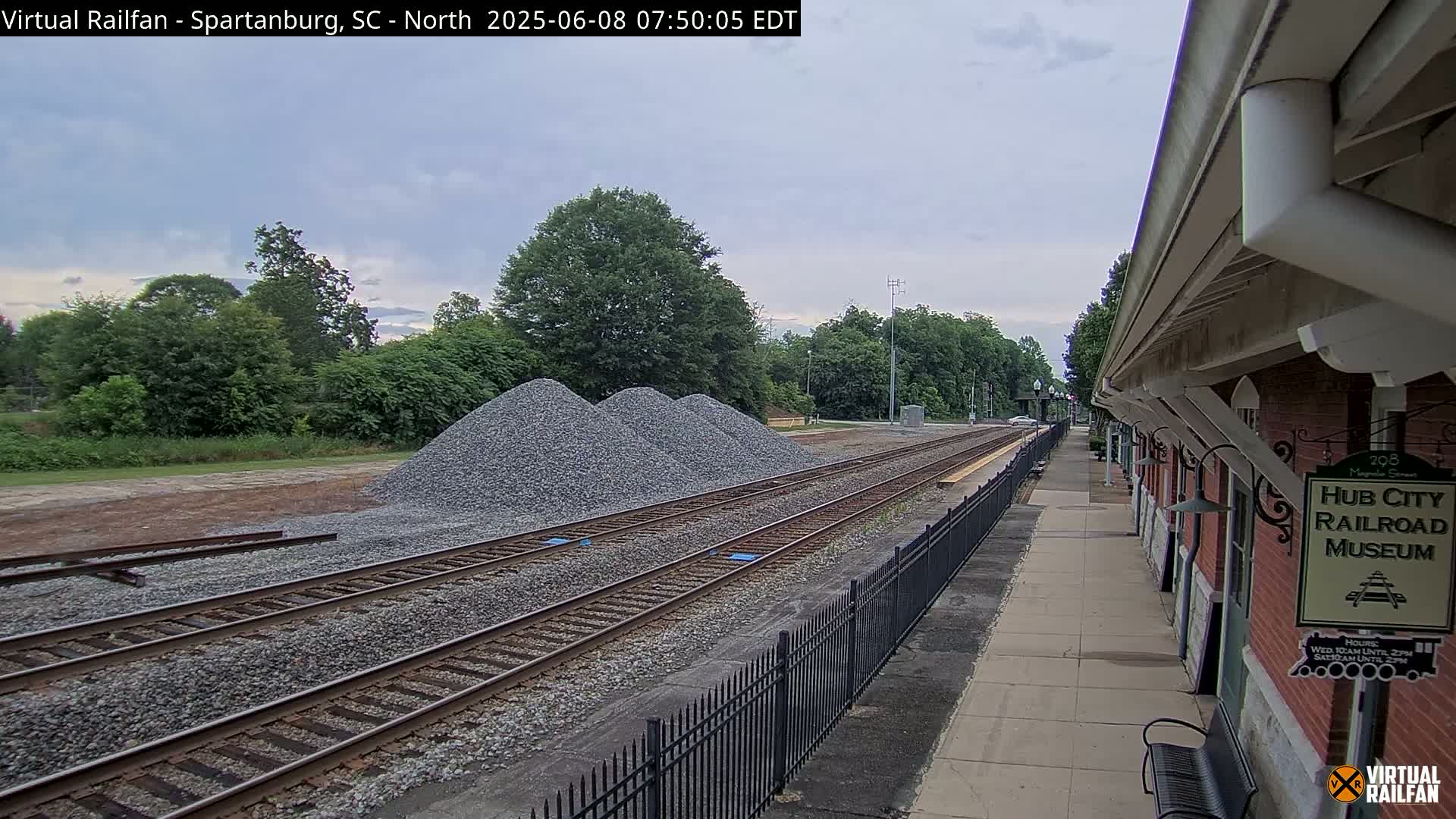 A railway platform next to a brick building stretches alongside train tracks, with piles of gravel nearby under an overcast sky.
