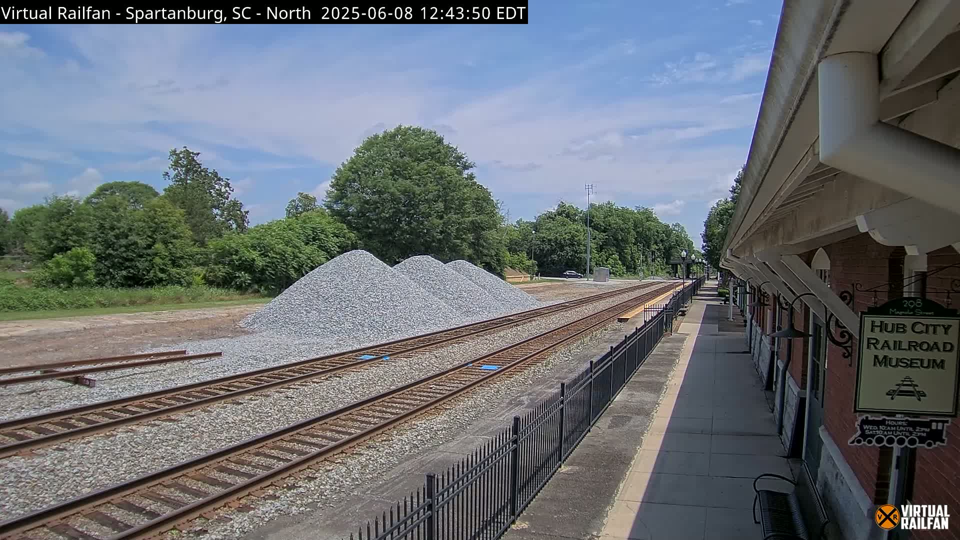 Under a partly cloudy sky, railroad tracks extend from a train platform past several piles of gravel toward a line of trees.