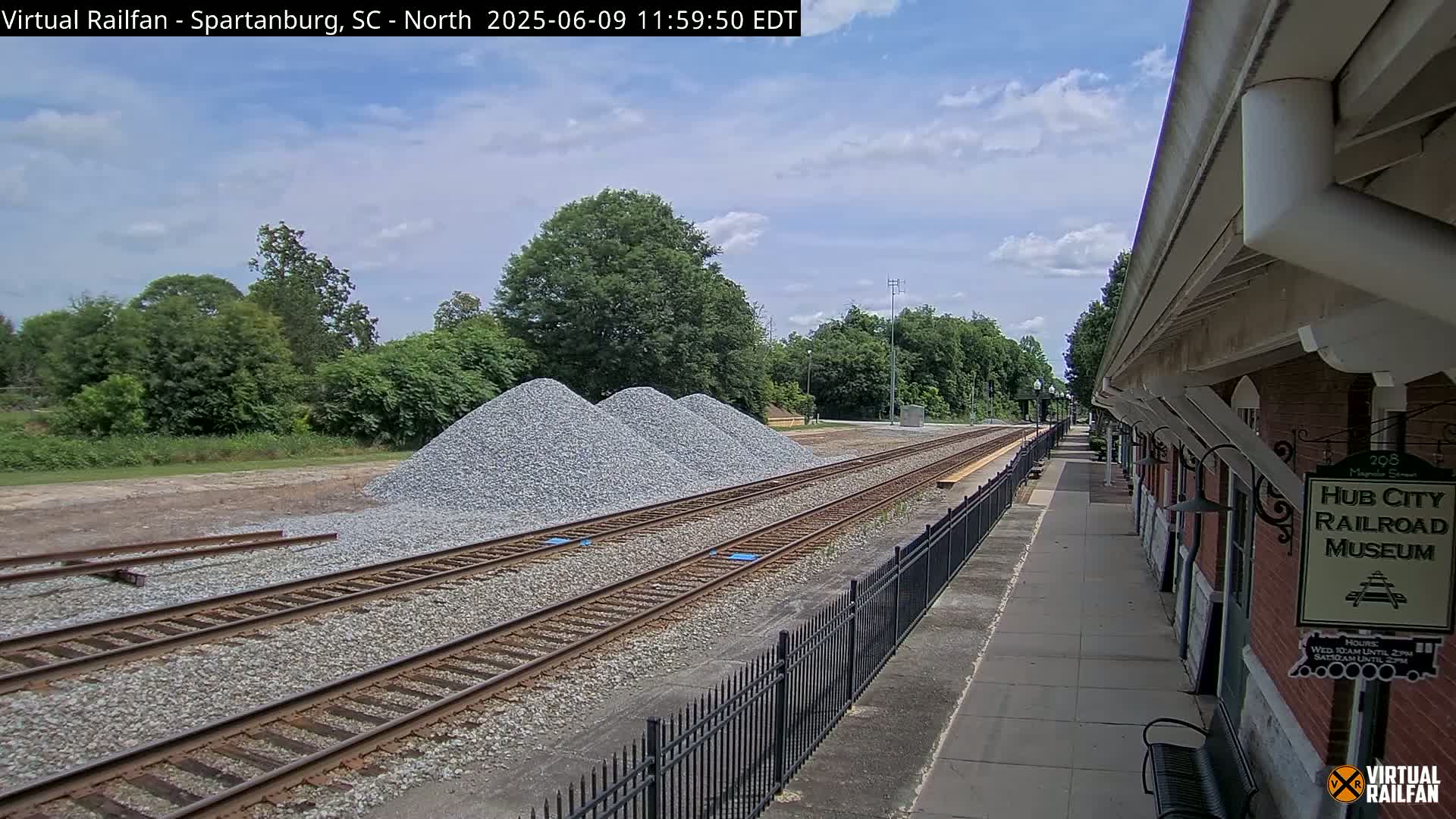 Under a partly cloudy sky, railroad tracks extend from a train platform beside a brick building toward piles of gravel.