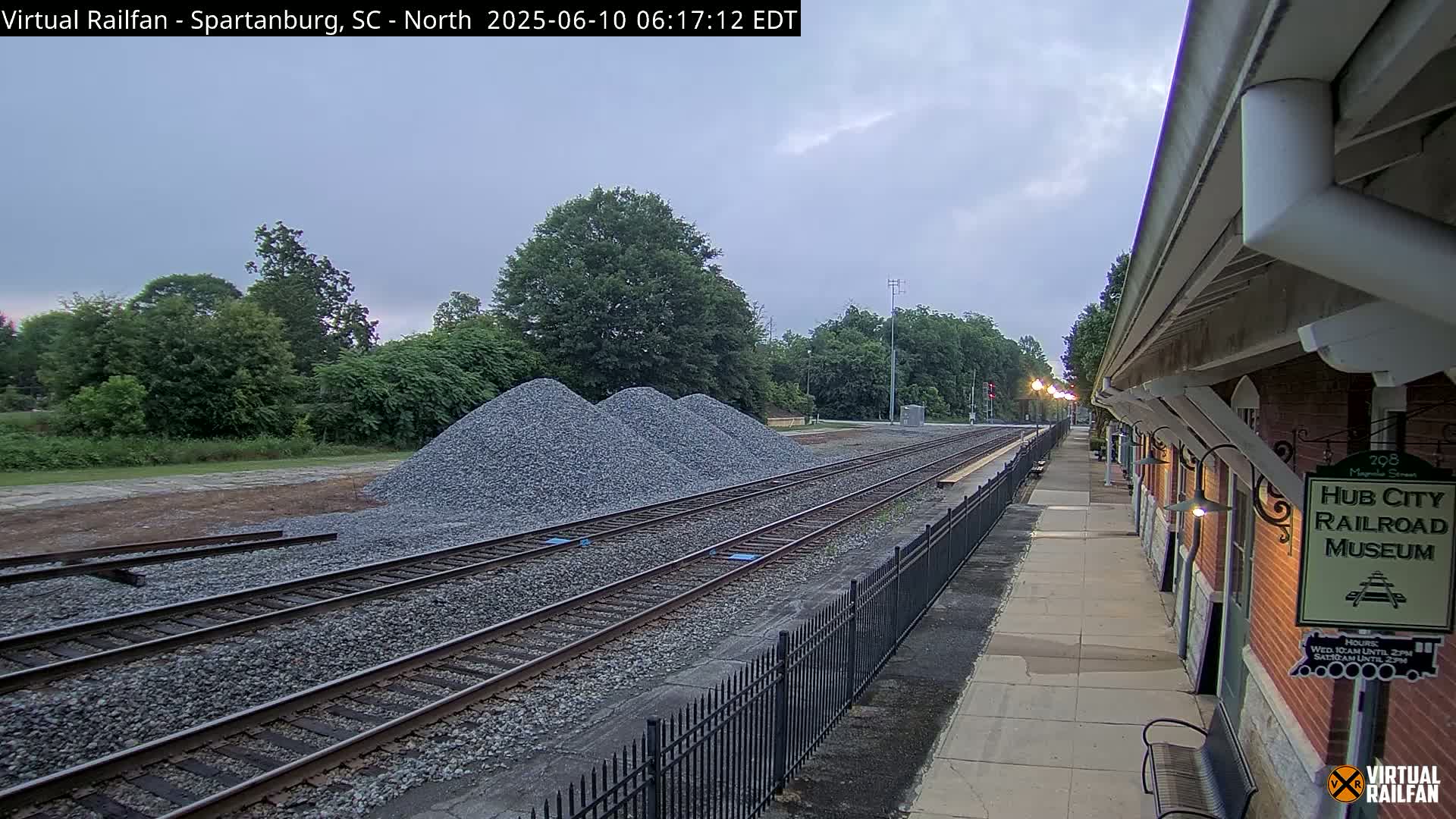 Train tracks and a platform next to a brick building are visible under an overcast sky, with piles of gravel nearby.