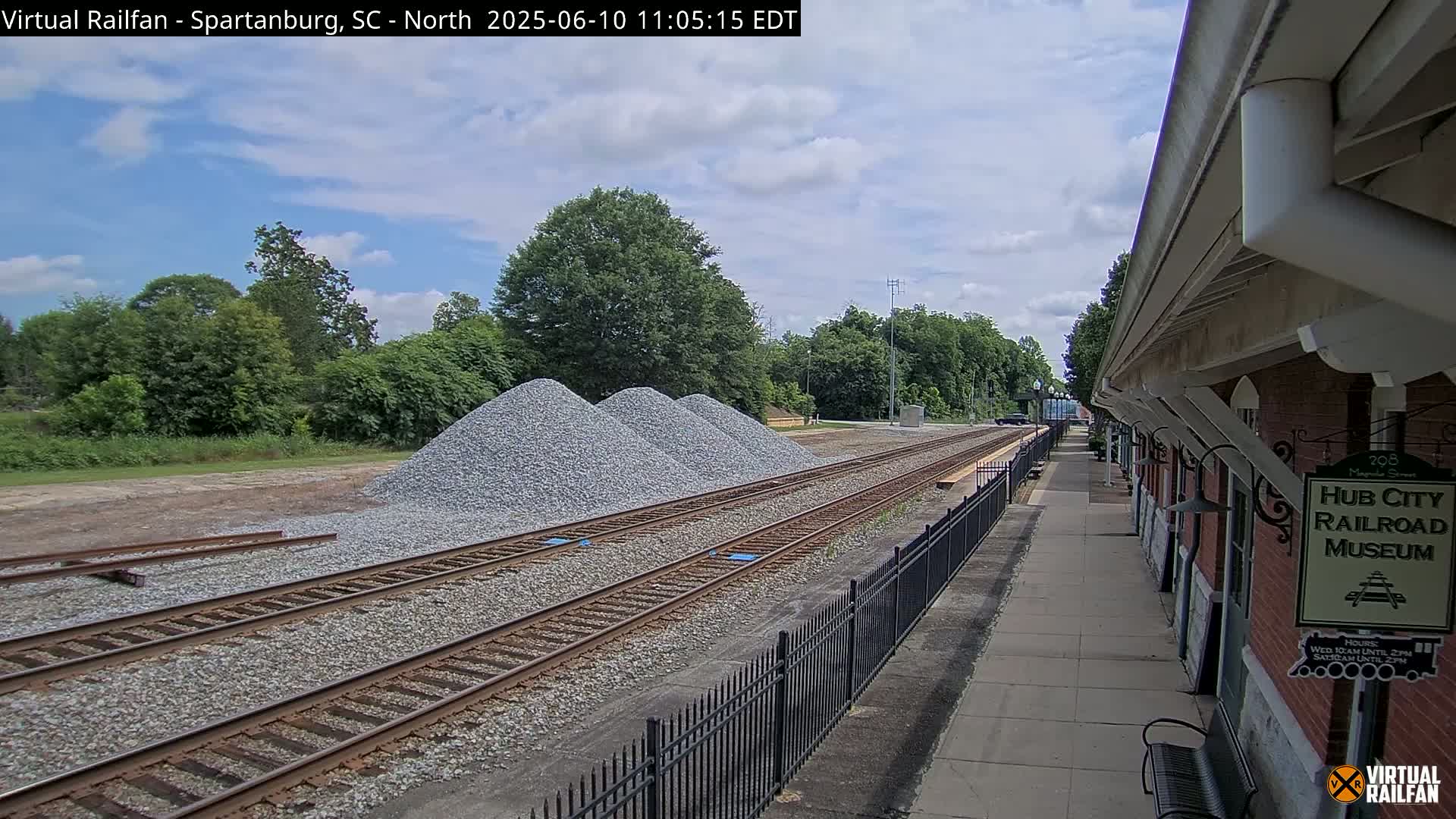 Under a partly cloudy sky, railroad tracks run alongside a brick building next to piles of gravel.