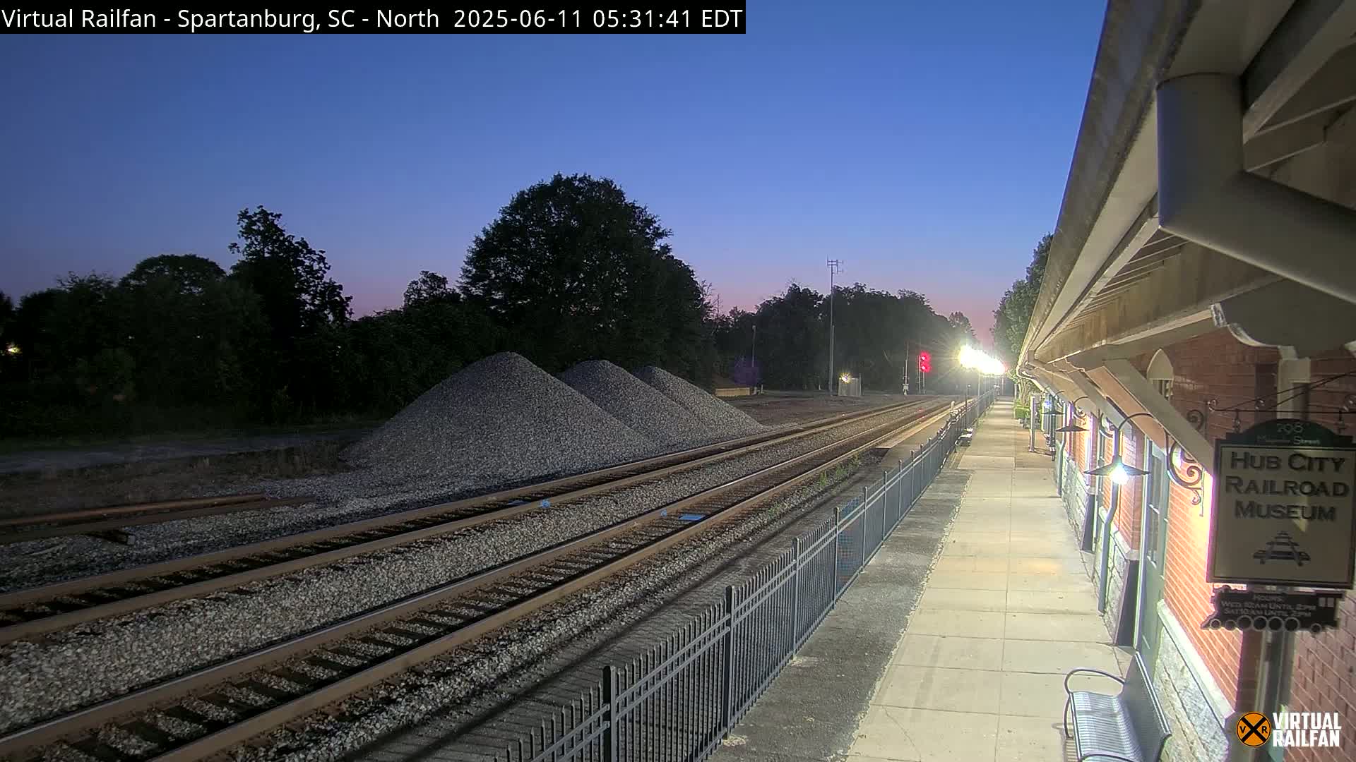 Under a twilight sky, railroad tracks extend from a train platform toward piles of gravel, bordered by a fence and trees.
