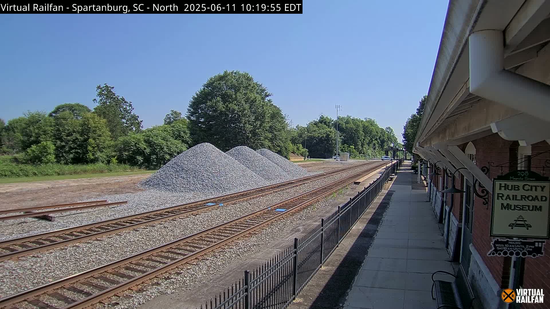 Under a clear blue sky, railroad tracks extend from a train depot past piles of gravel toward a line of trees.