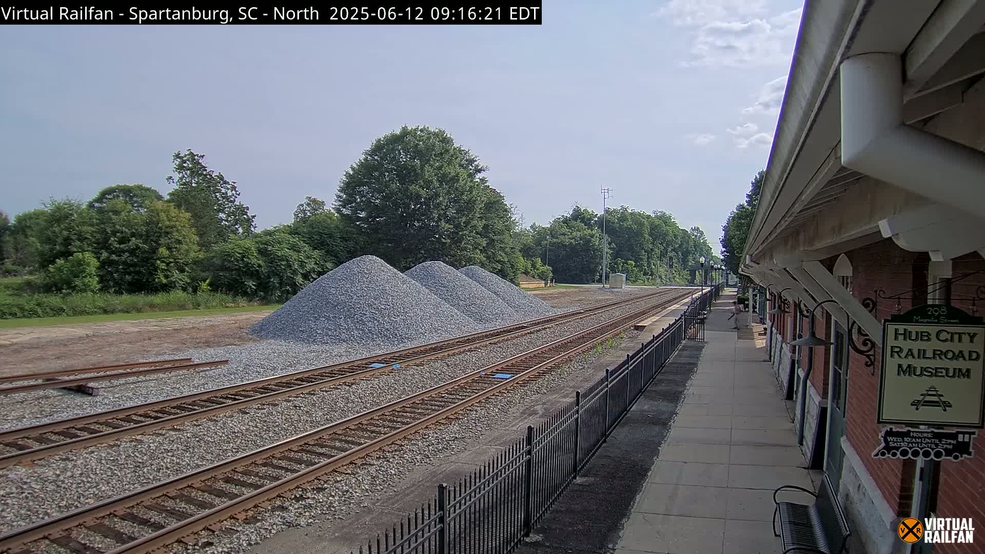 A partly sunny day reveals train tracks with several piles of gravel beside them, near a brick railroad museum and platform.