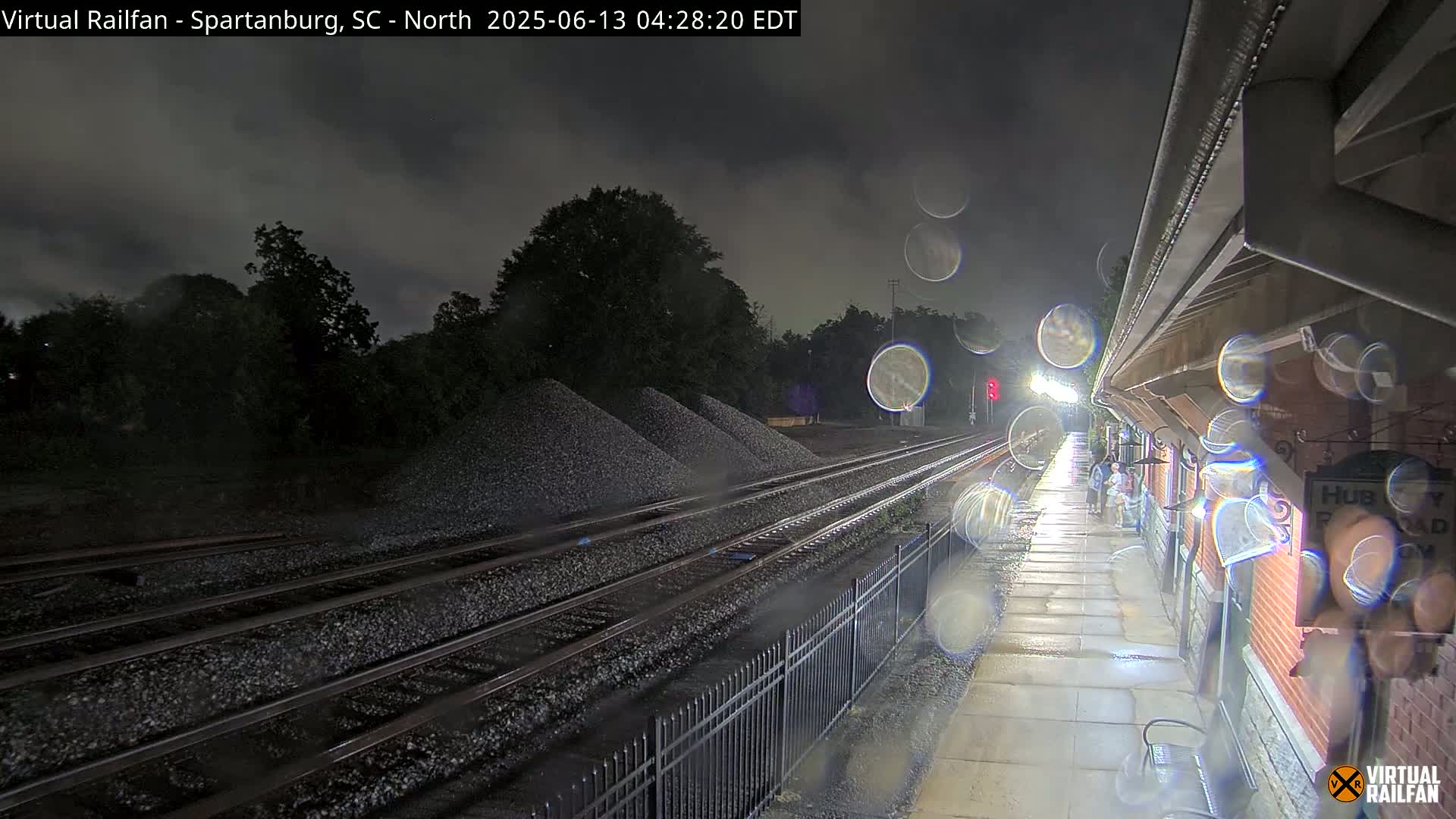 A nighttime view of rain-slicked train tracks and a partially visible train station platform under a dark, cloudy sky.