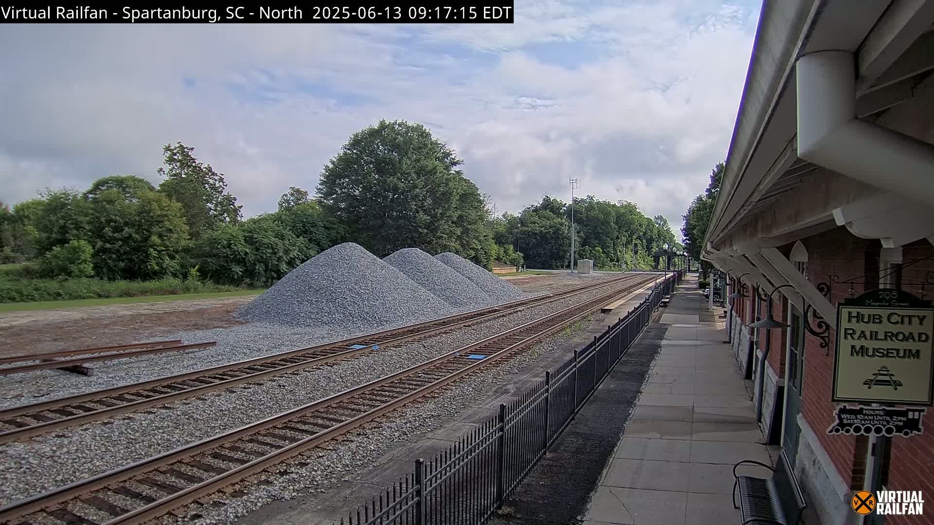 A partly cloudy day at a railroad station with multiple piles of gravel beside train tracks, next to a brick building.
