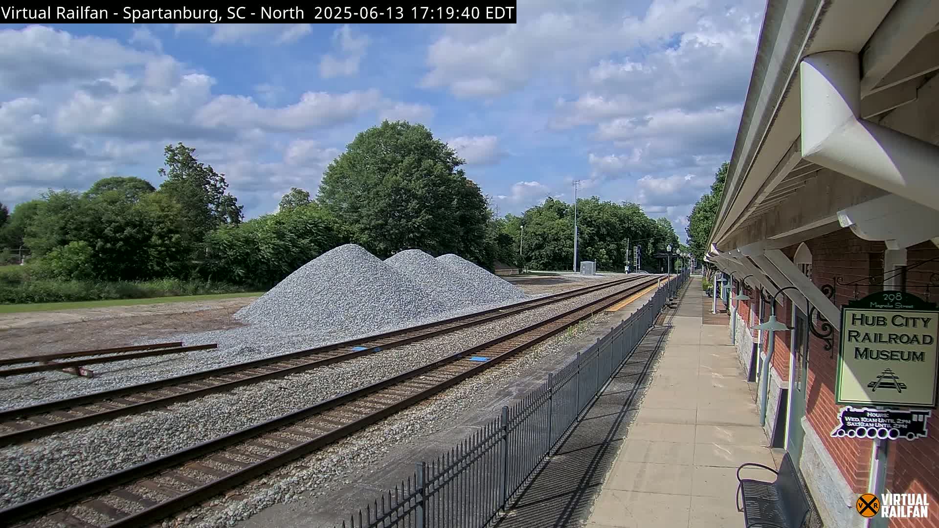 A partly cloudy day shows railroad tracks next to a brick building, with piles of gravel nearby.
