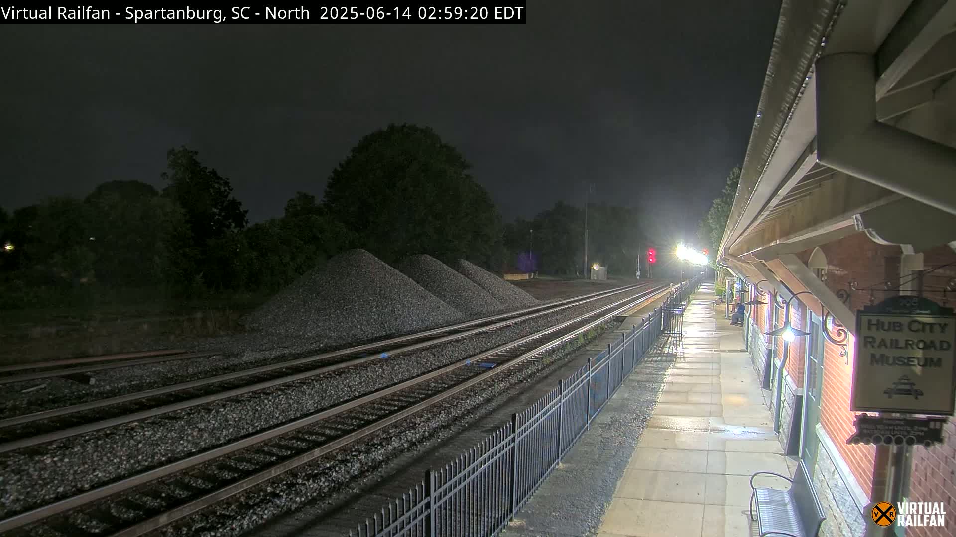 A partly cloudy day shows railroad tracks next to a brick building, with piles of gravel nearby.
