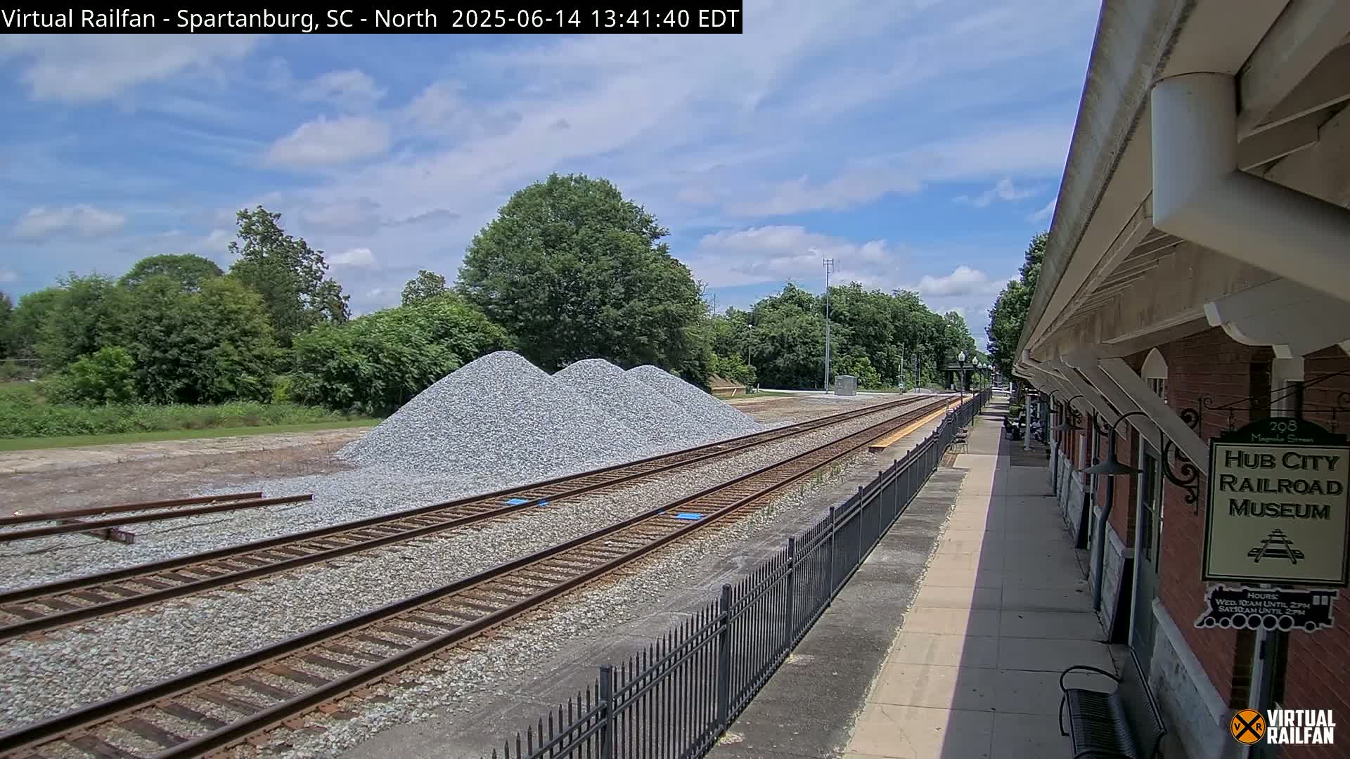Under a partly cloudy sky, railroad tracks extend from a platform next to a brick building, with piles of gravel nearby.
