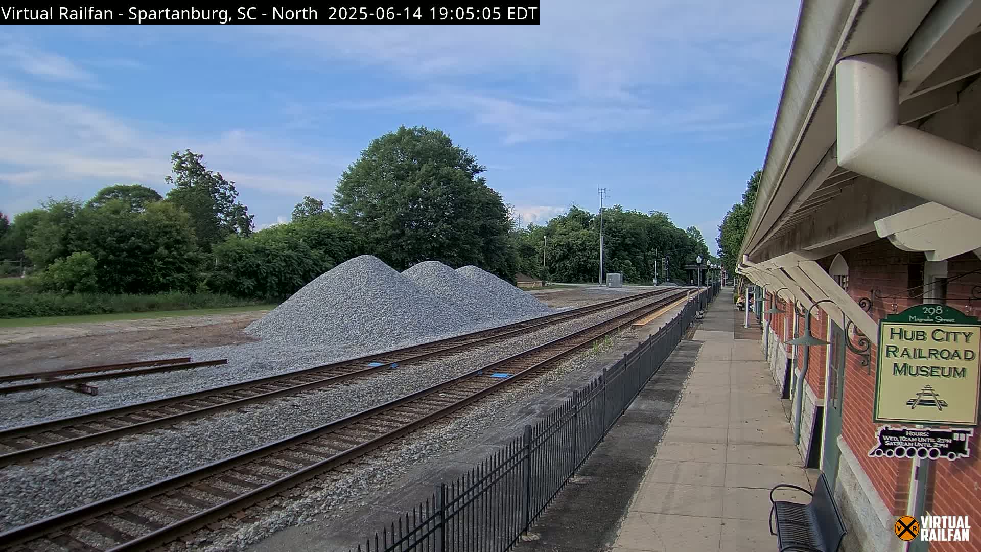 Under a partly cloudy sky, railroad tracks extend from a platform next to a brick building, with piles of gravel nearby.