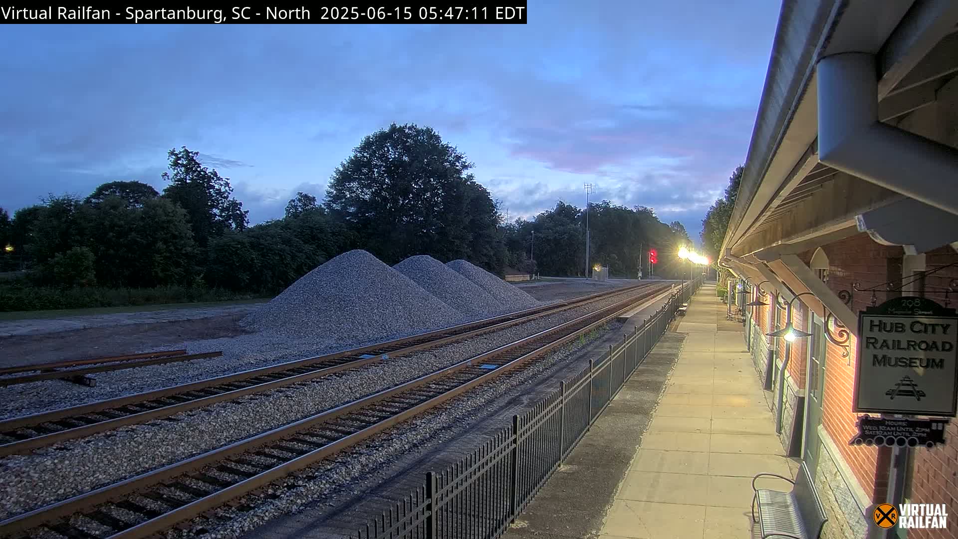 Under a twilight sky, railroad tracks run alongside a brick building and a paved walkway, with piles of gravel nearby.