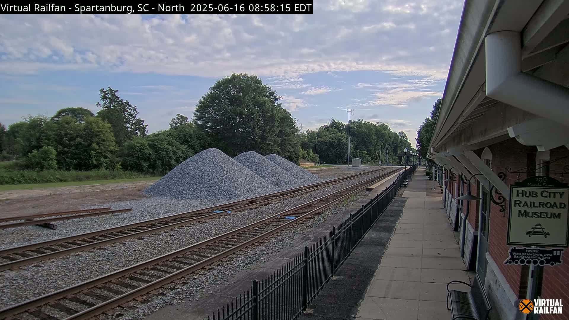 Several piles of gravel are situated next to a railway track beside a brick building under a partly cloudy sky.