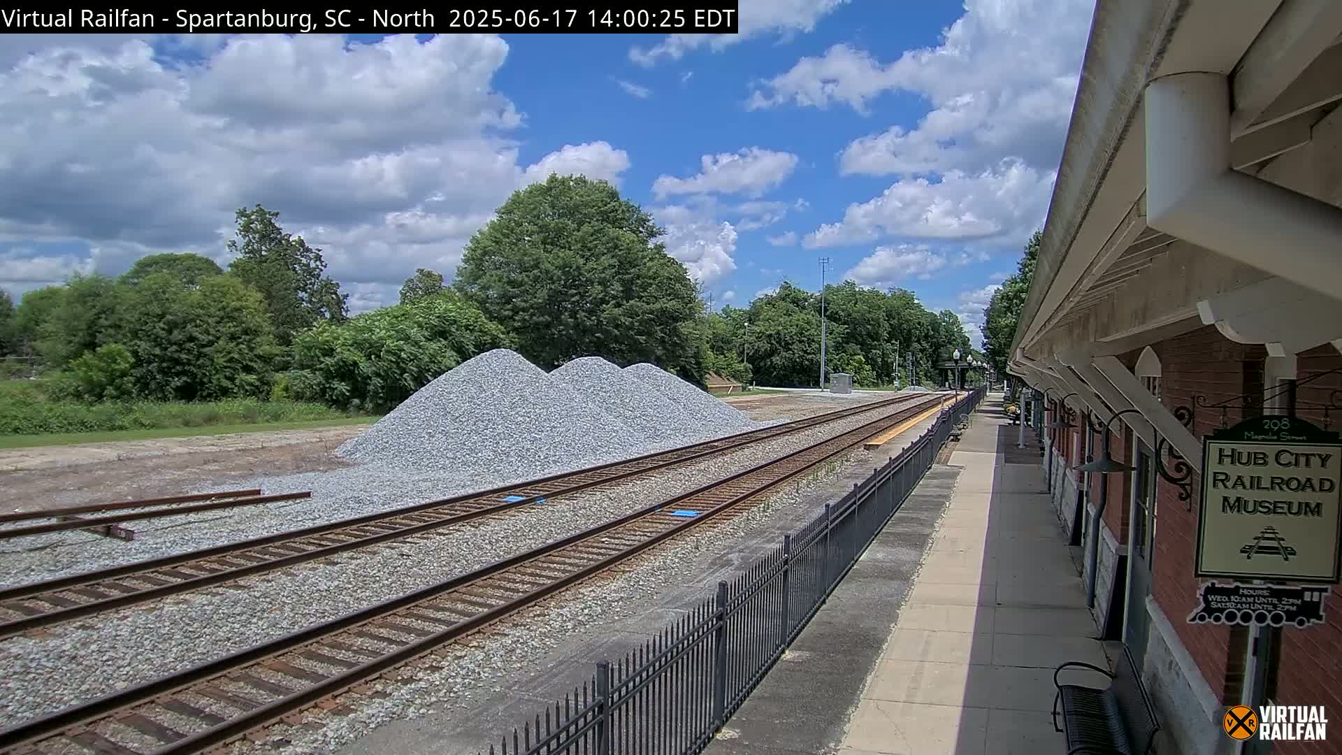 A partly sunny day shows a railroad track leading to a brick building next to a pile of gravel, with lush green trees in the background.