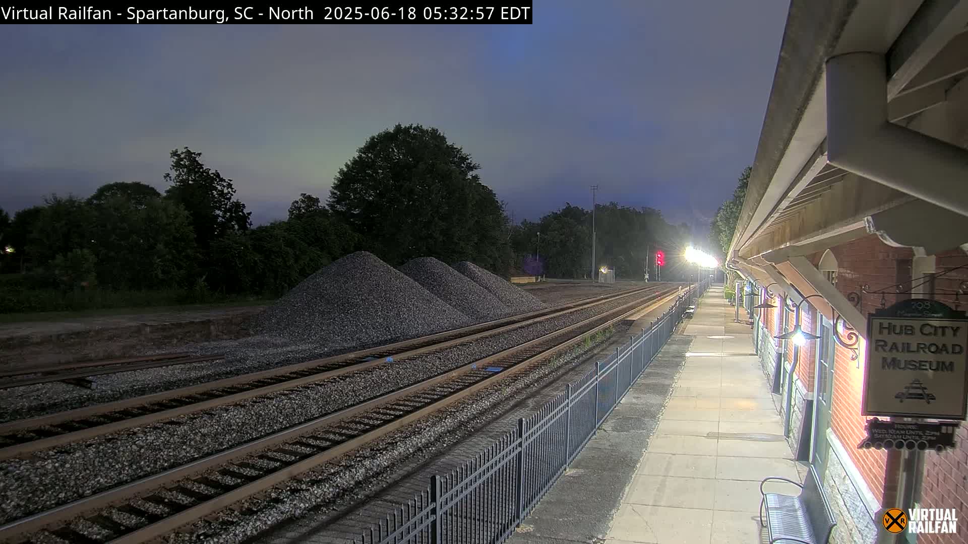 Under a twilight sky, railroad tracks extend toward a pile of gravel near a brick building with exterior lights.
