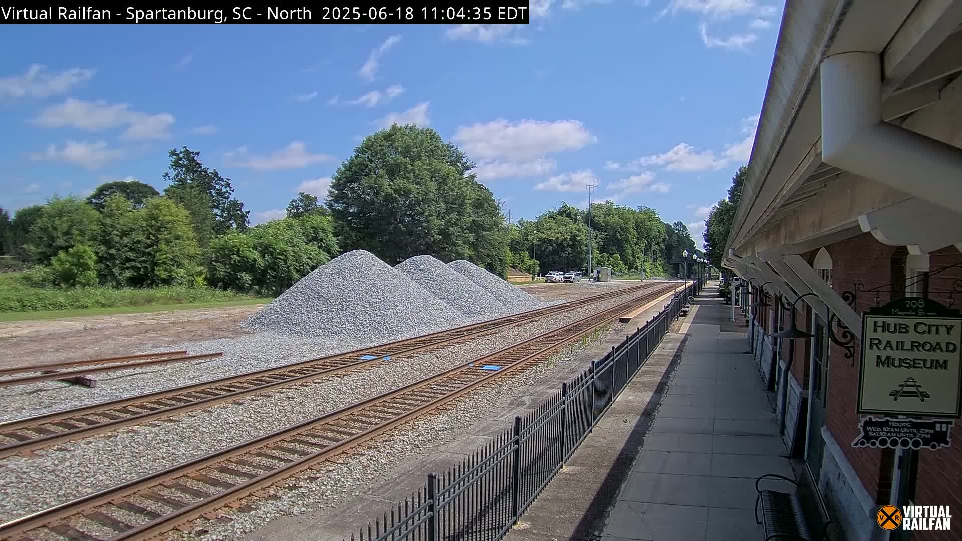 Under a partly cloudy sky, railroad tracks extend alongside a brick building and several piles of gravel.