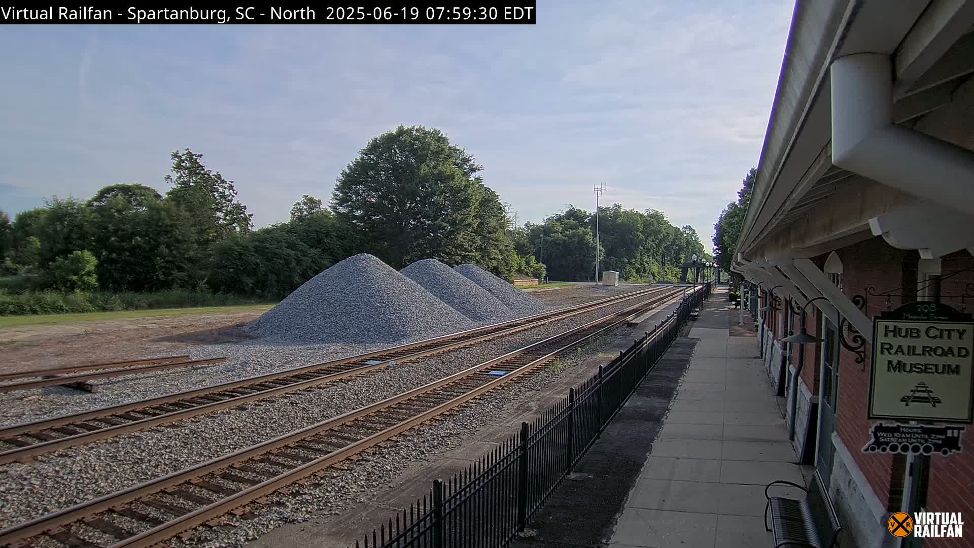 Three piles of gravel sit beside railroad tracks next to a train station platform on a partly cloudy day.