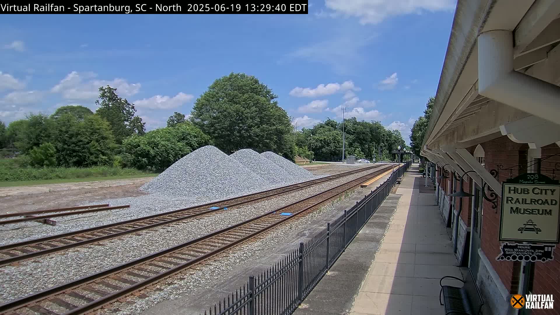 A partly cloudy day reveals train tracks, a section of a brick building, and several piles of gravel next to lush green trees.