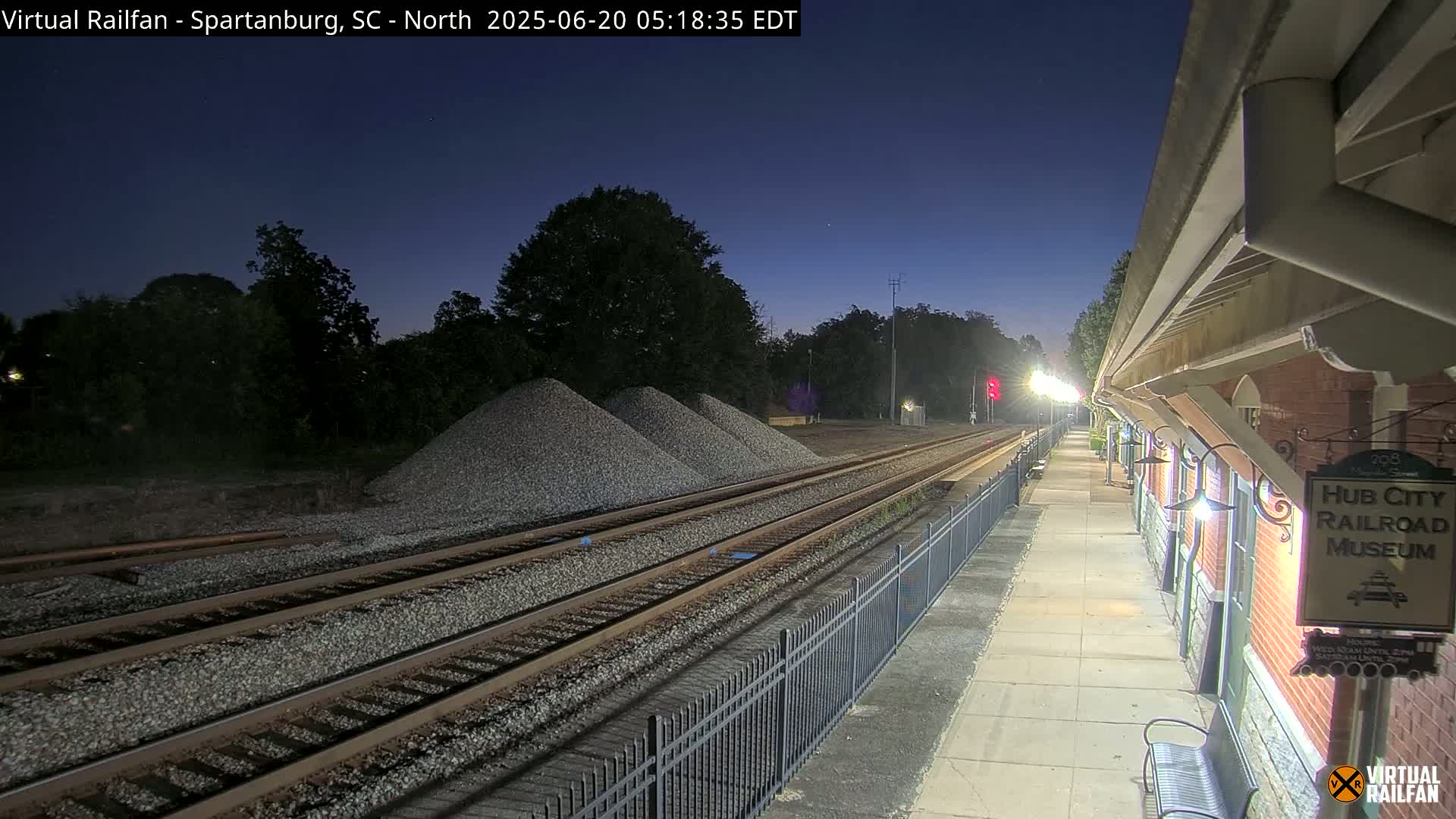 Under a twilight sky, railroad tracks extend from a nighttime train platform toward piles of gravel and trees.