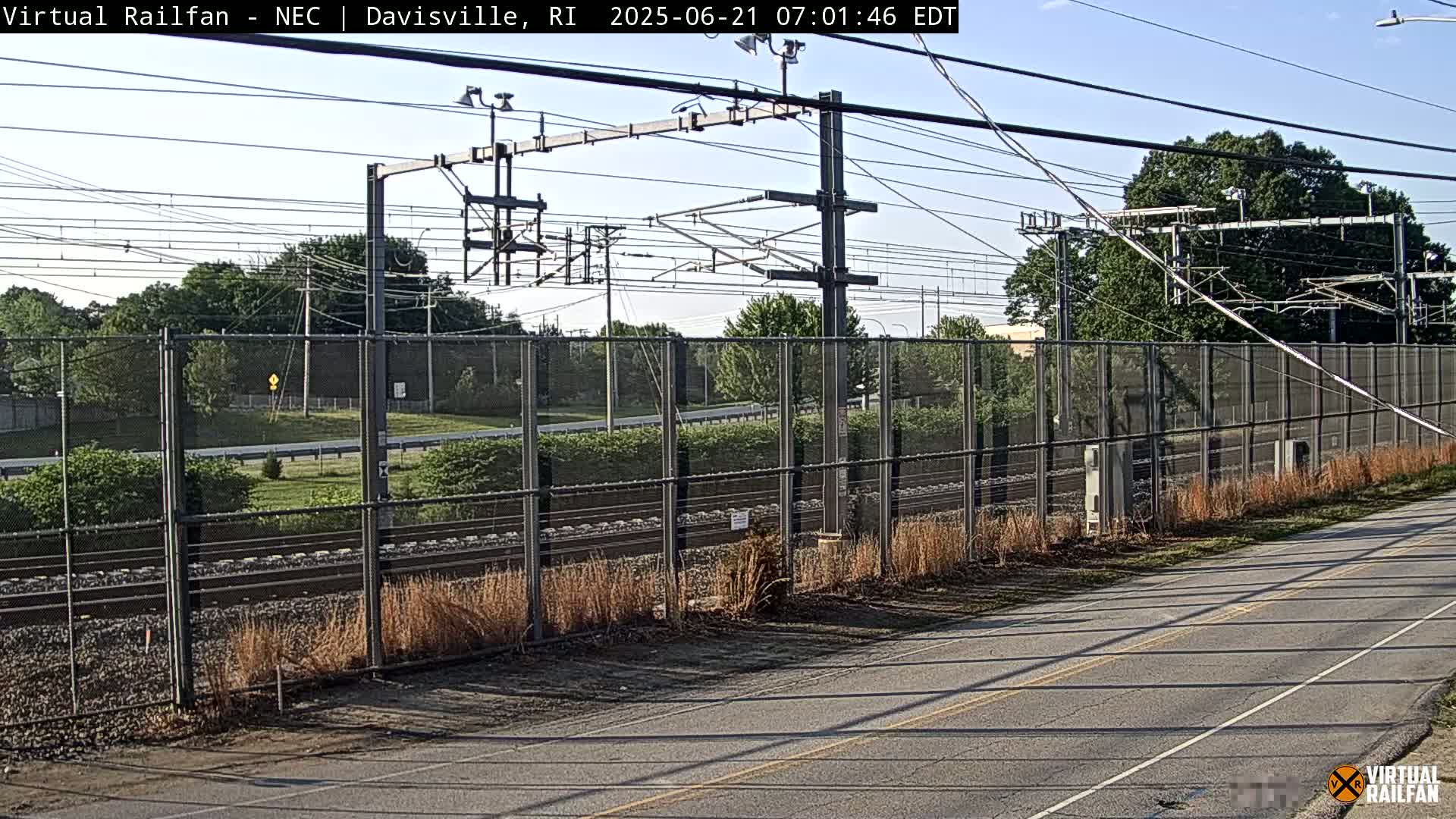 A paved road runs alongside a chain-link fence enclosing railroad tracks under a mostly clear sky.